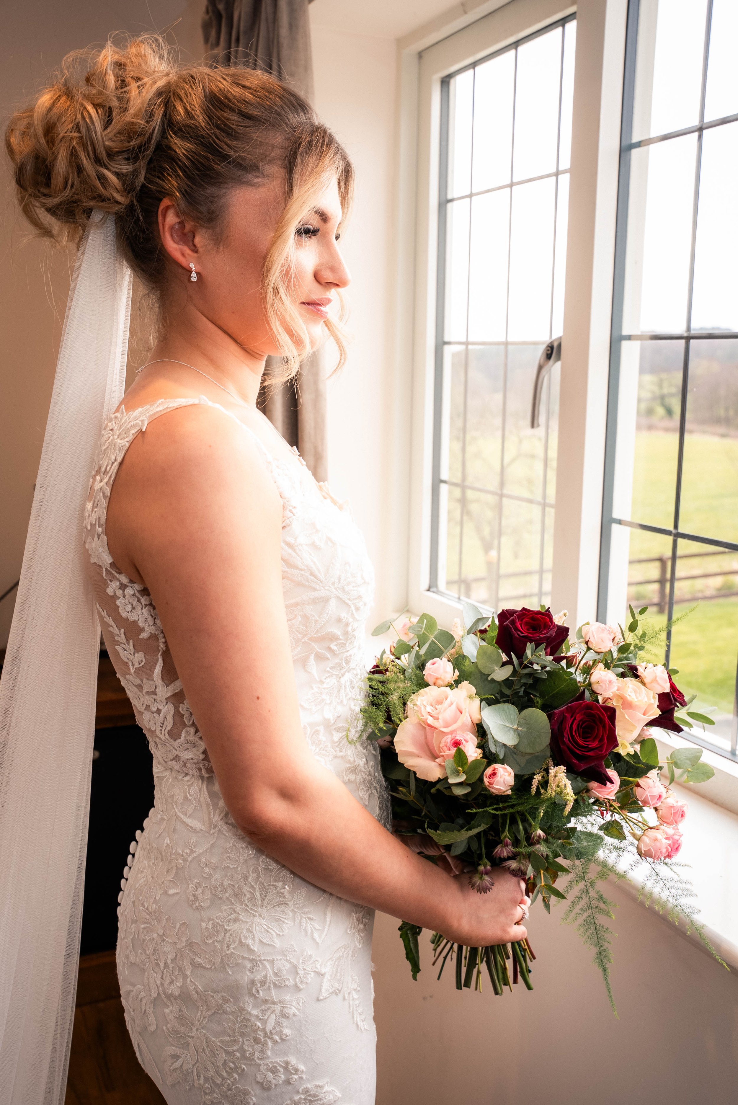 A bride in a lace wedding dress standing by a window holding a bouquet of roses and greenery, with a peaceful outdoor view in the background.