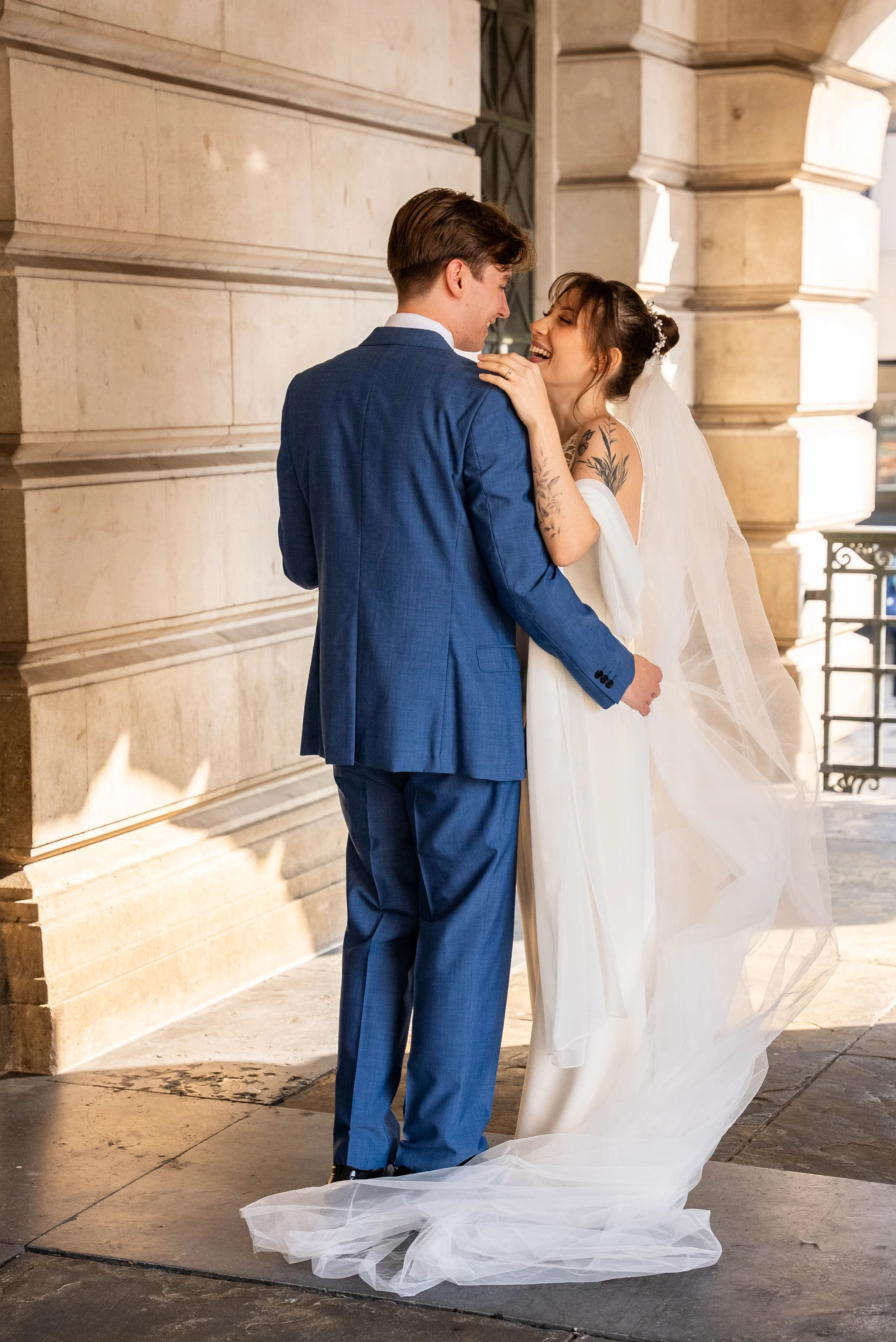 A newlywed couple shares a joyful moment on their wedding day, standing close to each other outside a stone building, with the bride wearing a white wedding gown and veil, and the groom in a blue suit.