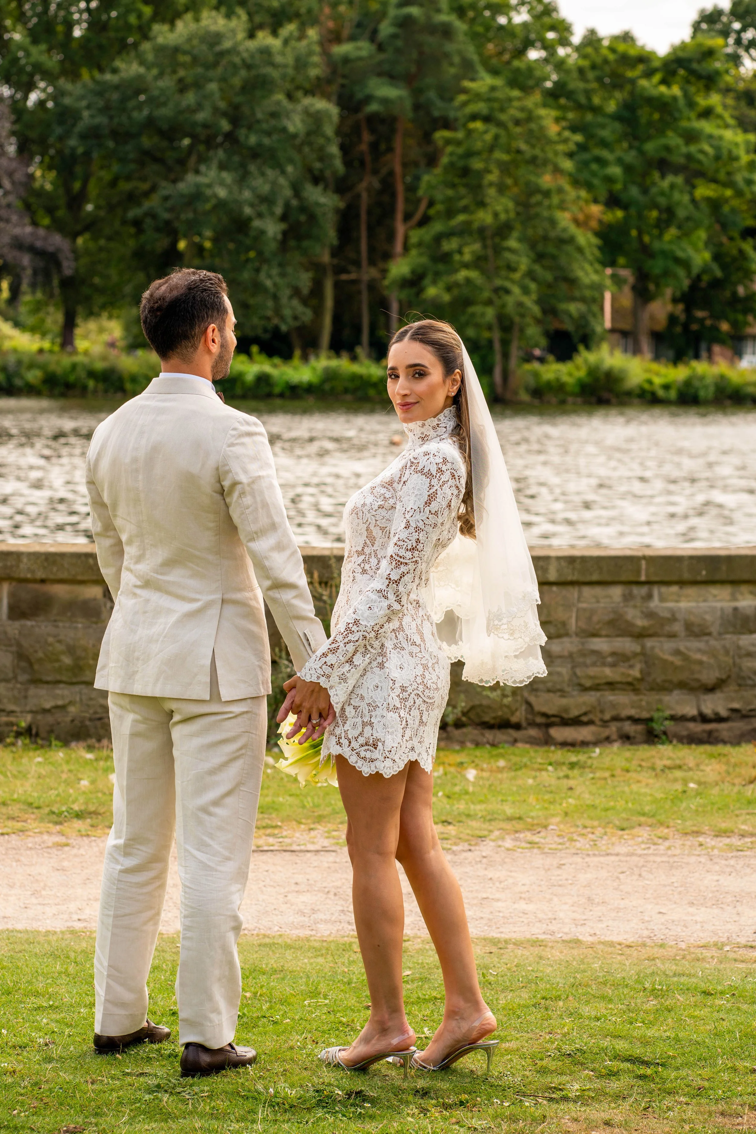 A bride and groom holding hands by a river, with trees and greenery in the background, during their wedding ceremony.