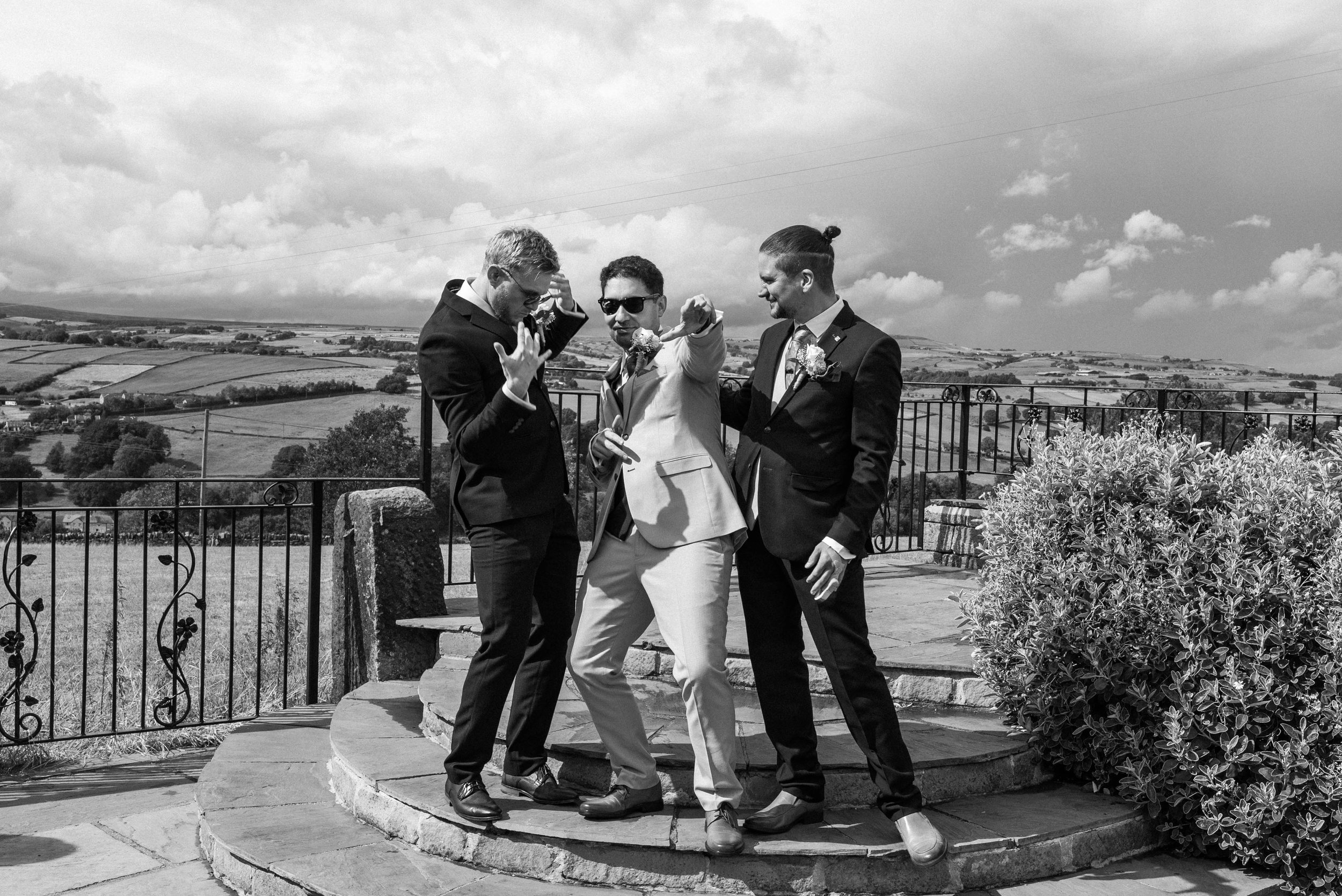 Three men in suits dancing and having fun on a stone platform outdoors, with a rural landscape and cloudy sky in the background.