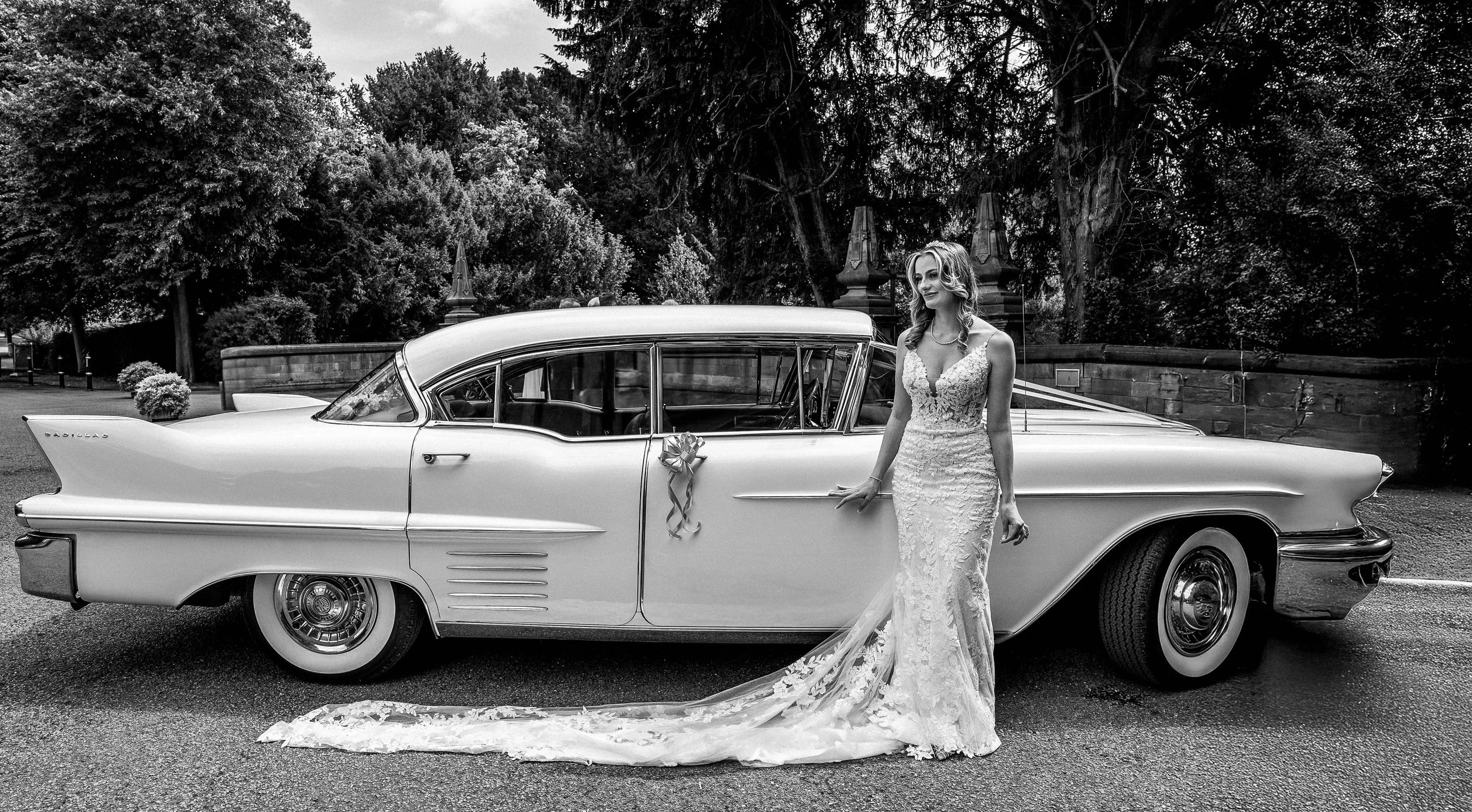 Black and white photo of a bride in a lace wedding dress standing beside a vintage car decorated with a ribbon, outdoors in a park with trees and a stone fence in the background.