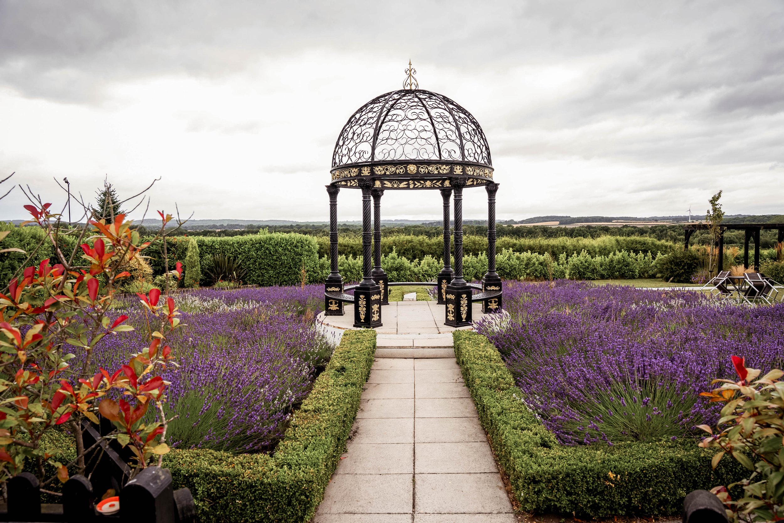 A garden with a paved path leading to a decorative black and gold gazebo surrounded by purple lavender flowers, green bushes, and trees under a cloudy sky.