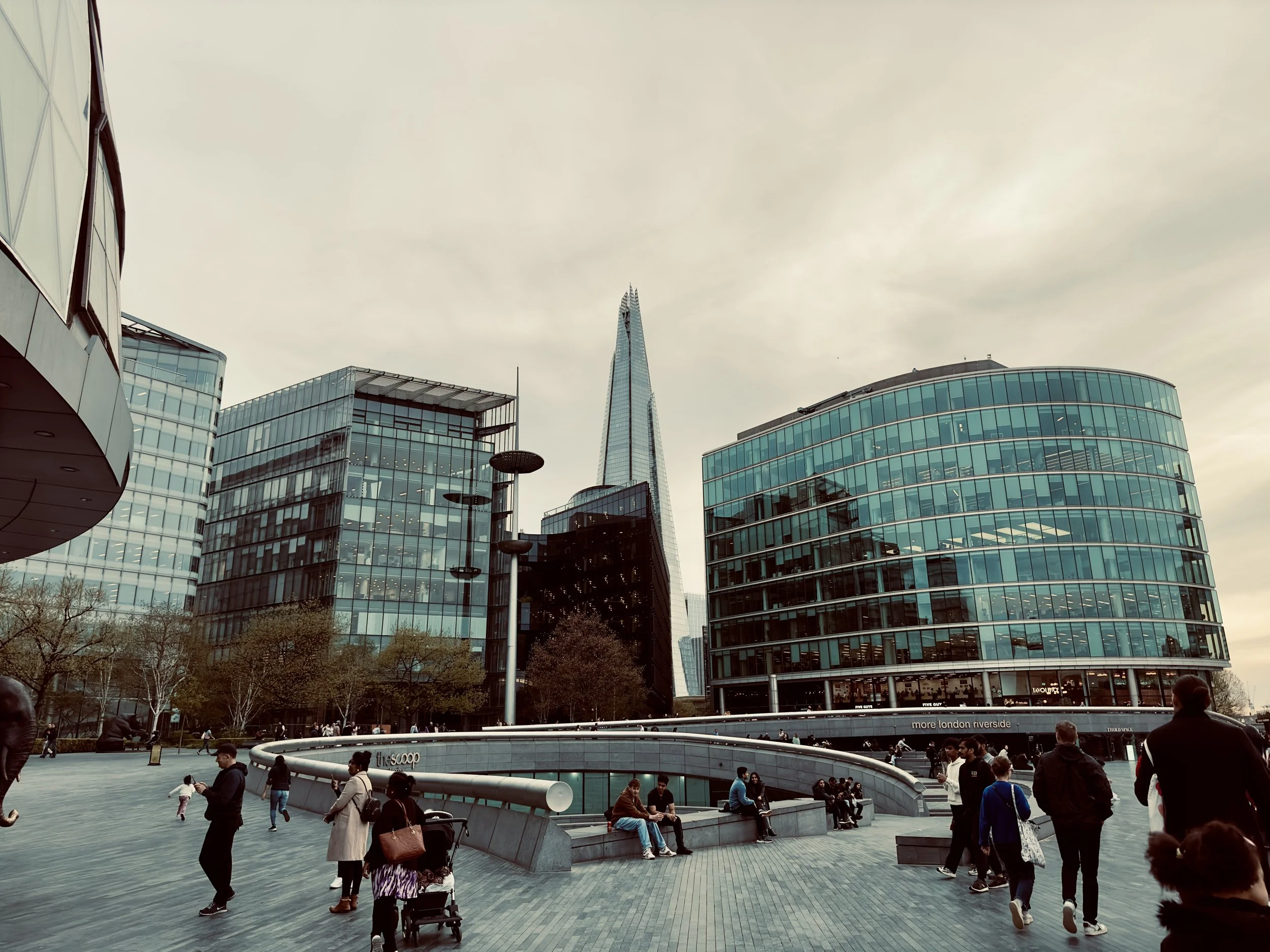 People walking and sitting in a modern urban plaza surrounded by glass office buildings and the tall, pointed Shard skyscraper in London.