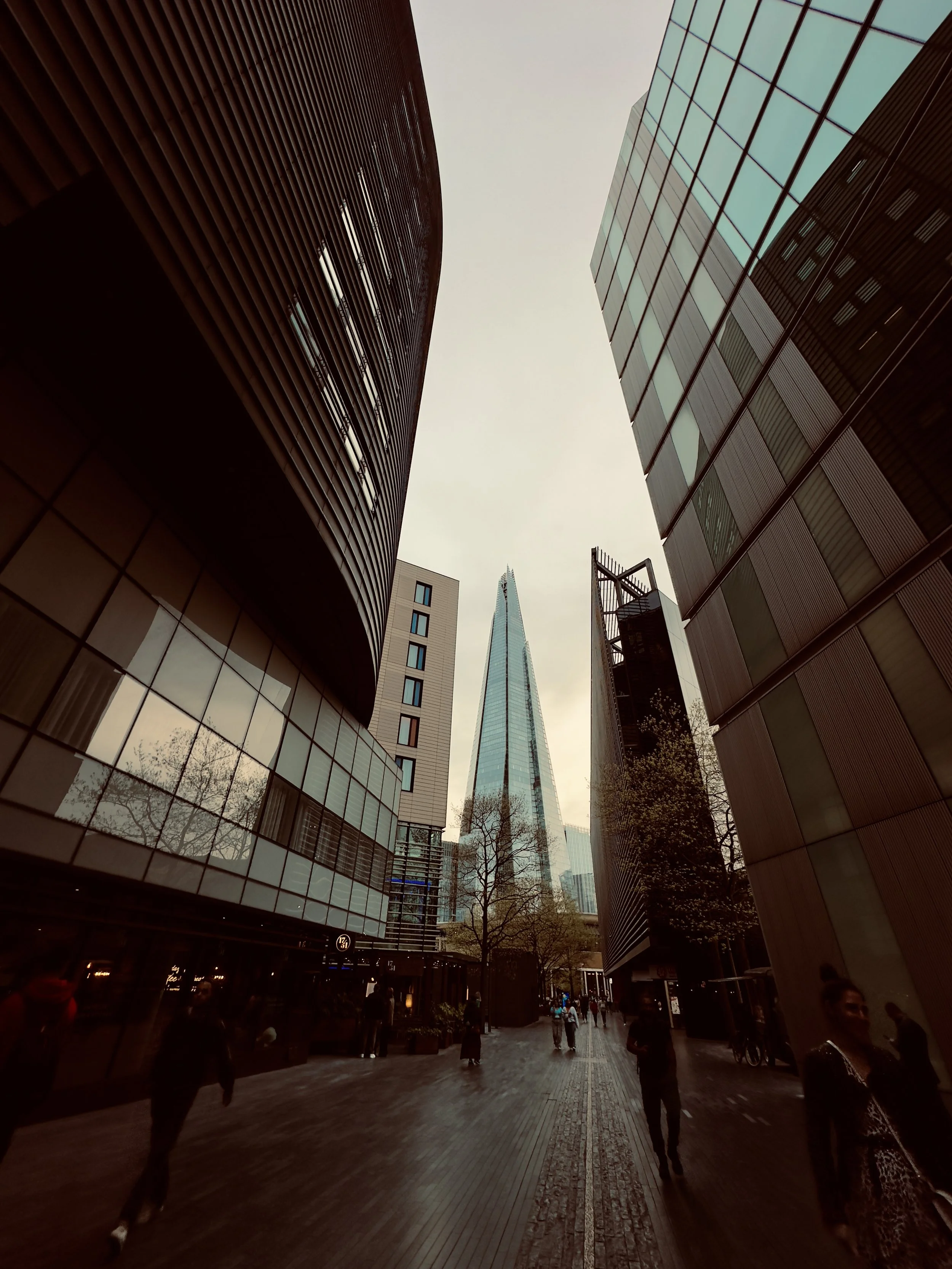 Tall glass skyscraper viewed from street level with surrounding modern buildings and pedestrians walking on wet pavement.