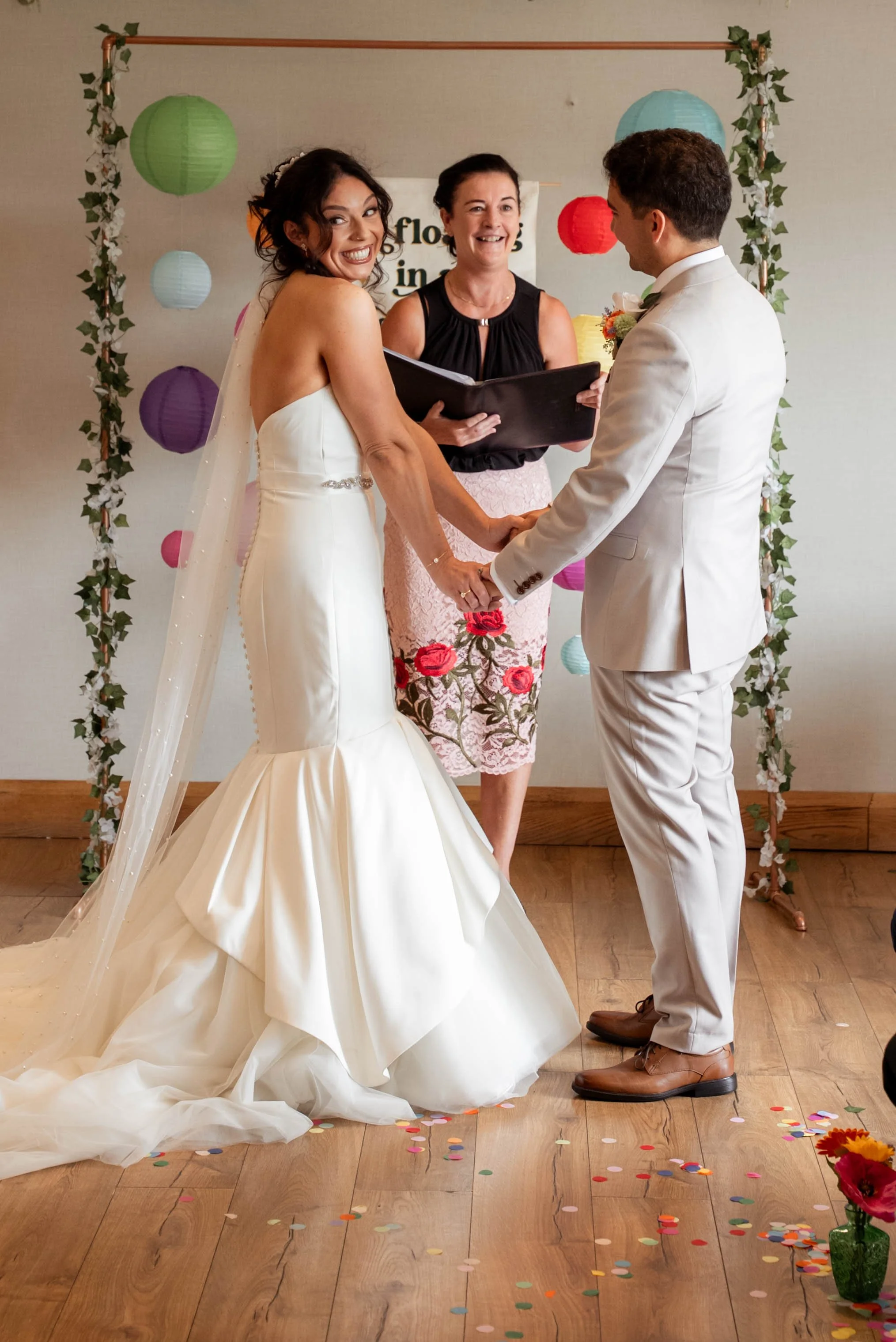 A bride and groom holding hands and smiling during their wedding ceremony, with an officiant standing behind them holding a book, decorated with colorful lanterns and confetti on the floor.