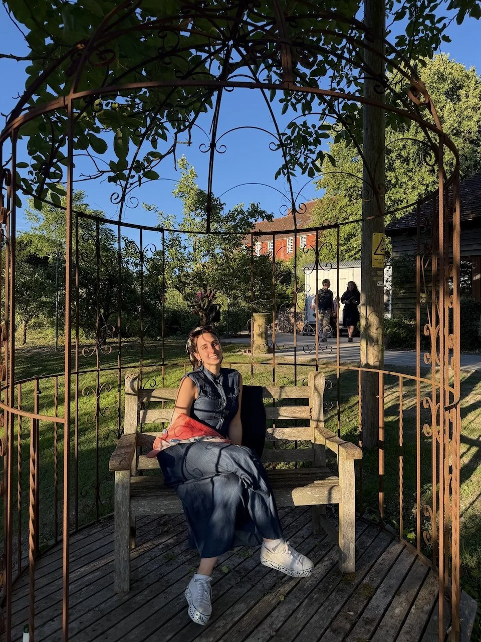 A young woman sitting on a wooden bench inside a decorative metal gazebo in a park, smiling at the camera during late afternoon sunlight. Behind her, trees, a house, and two people walking are visible.