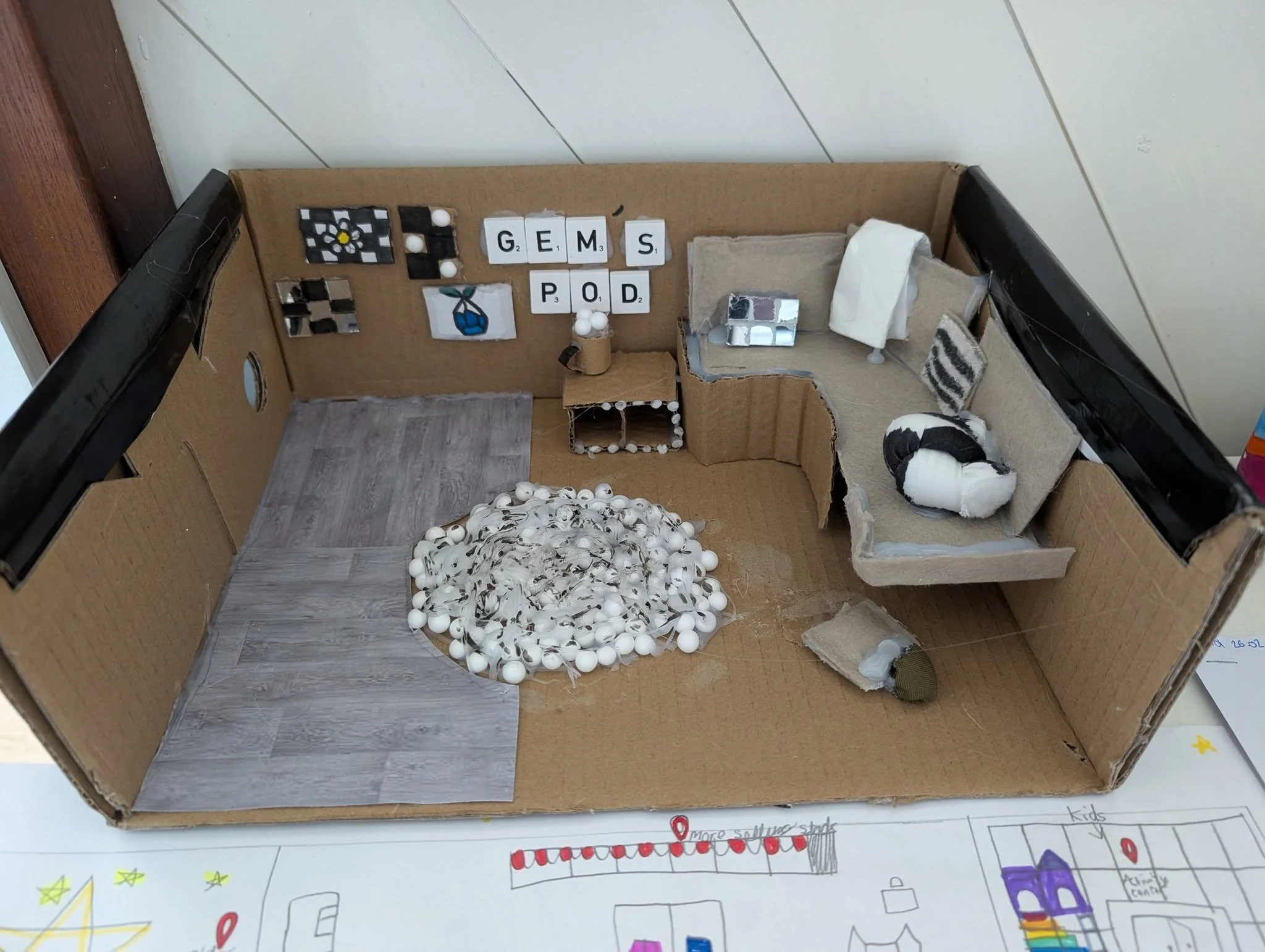 A miniature model of a living room built inside a cardboard box, with various small objects including a pile of white balls representing a fire, a black and white dog figure, a small sofa, and decorative items on the wall.