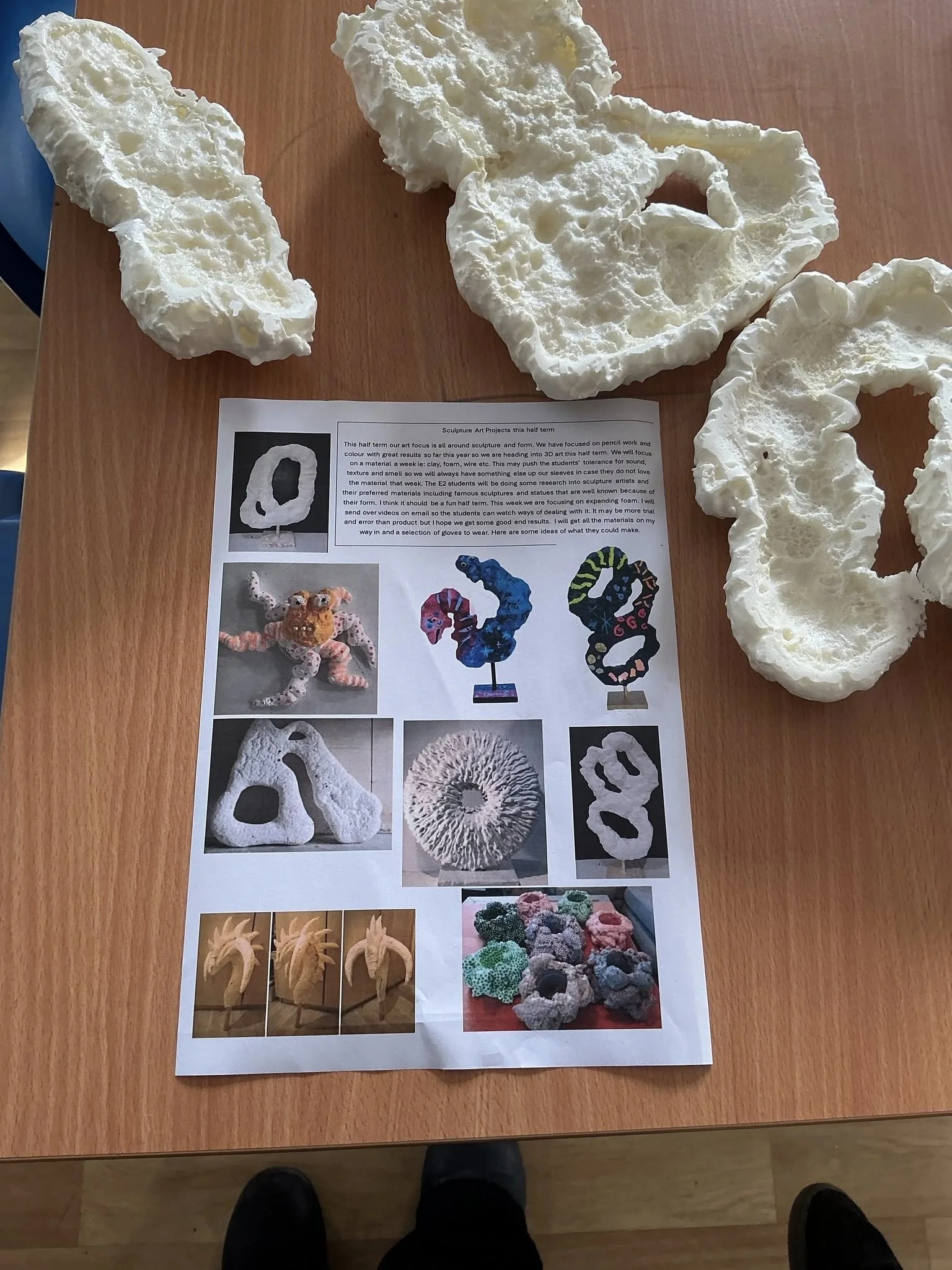 Sculpture art projects display on a wooden table, with various foam sculptures and a printed informational sheet featuring images of sculptures and project descriptions.