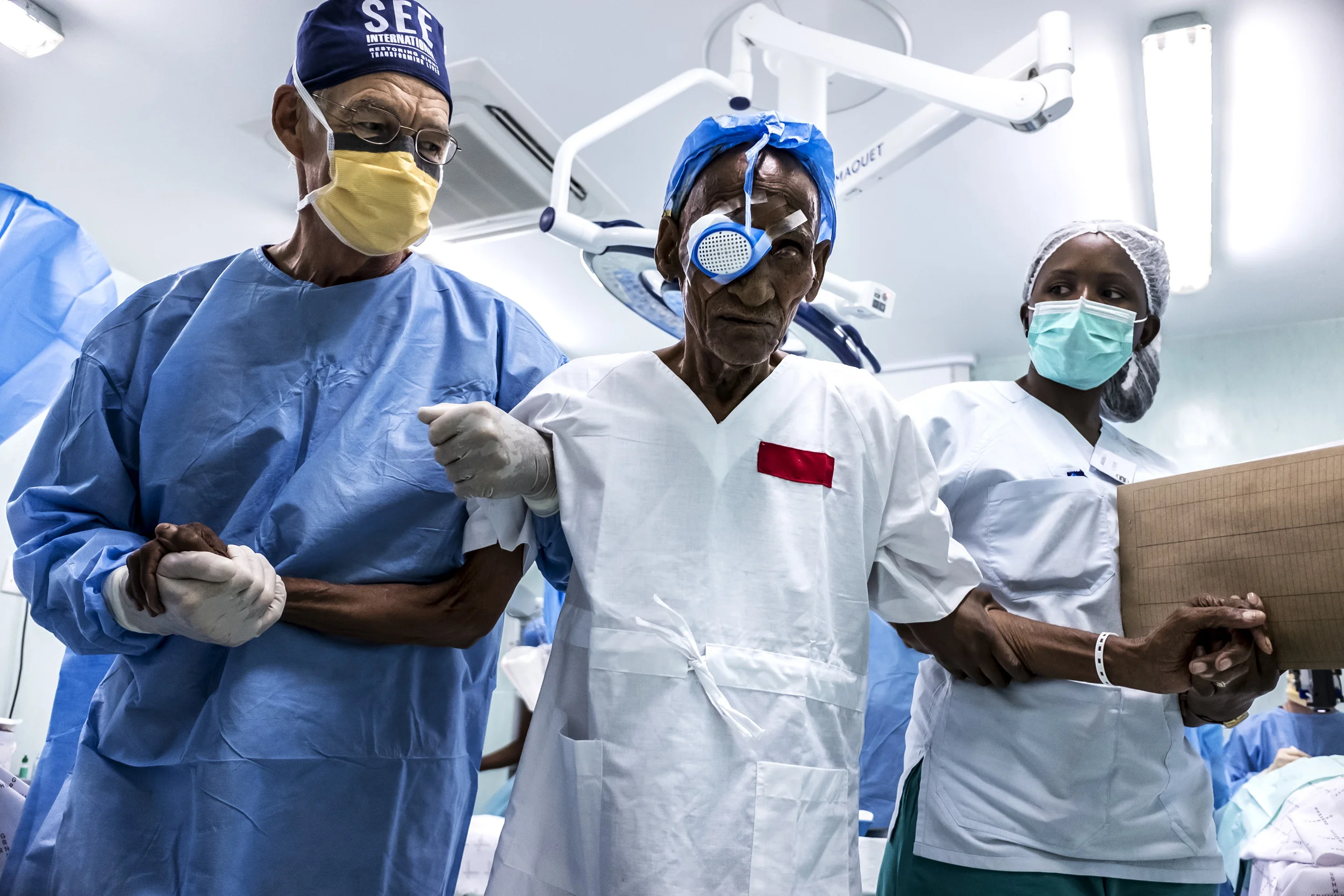 An eye camp for impoverished rural people on the Angolan/Namibian border run by SEE International and Mandela Award winner, Dr Helena Ndume. Surgeons are seen showing a 103 year old man back to his ward after successful eye surgery for cataracts.