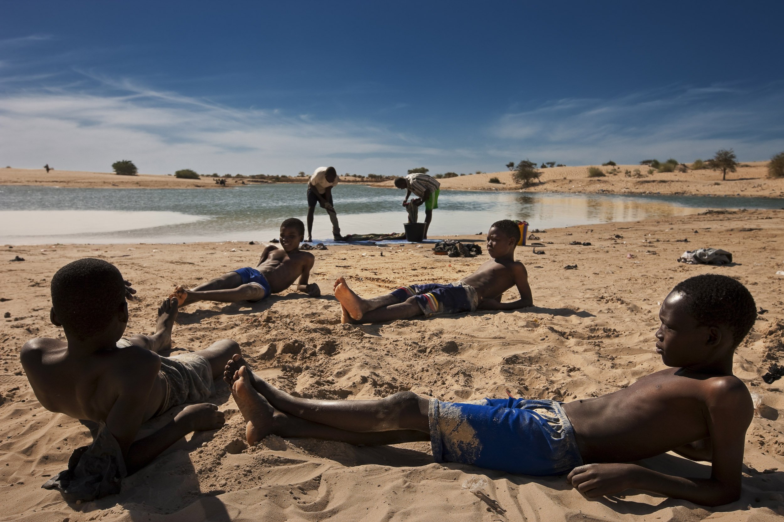 : TIMBUKTU, MALI, JANUARY 2010:Young boys lounge in the desert sand on the banks of the Libyan built Canal which brings river water to the heart of Timbuktu, Mali, 9 January 2010. In the background two young men clean their household carpet. The Can