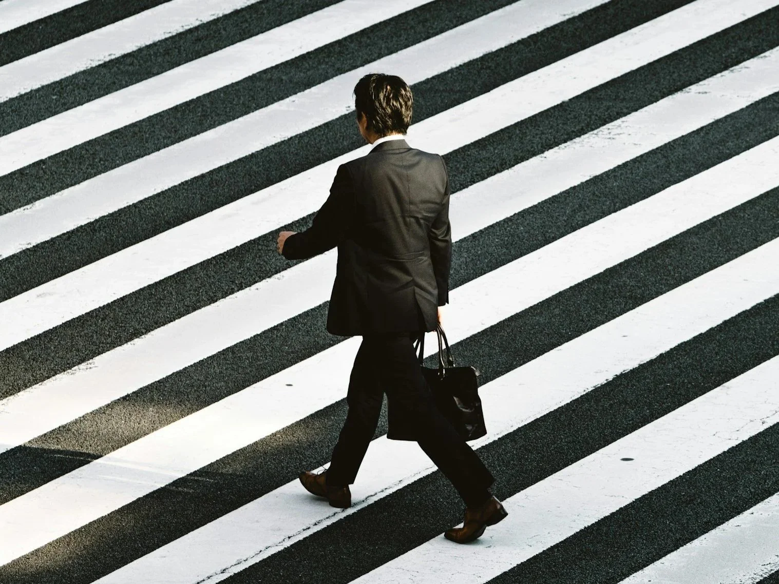 A person in formal business attire crossing a striped crosswalk in an urban setting.