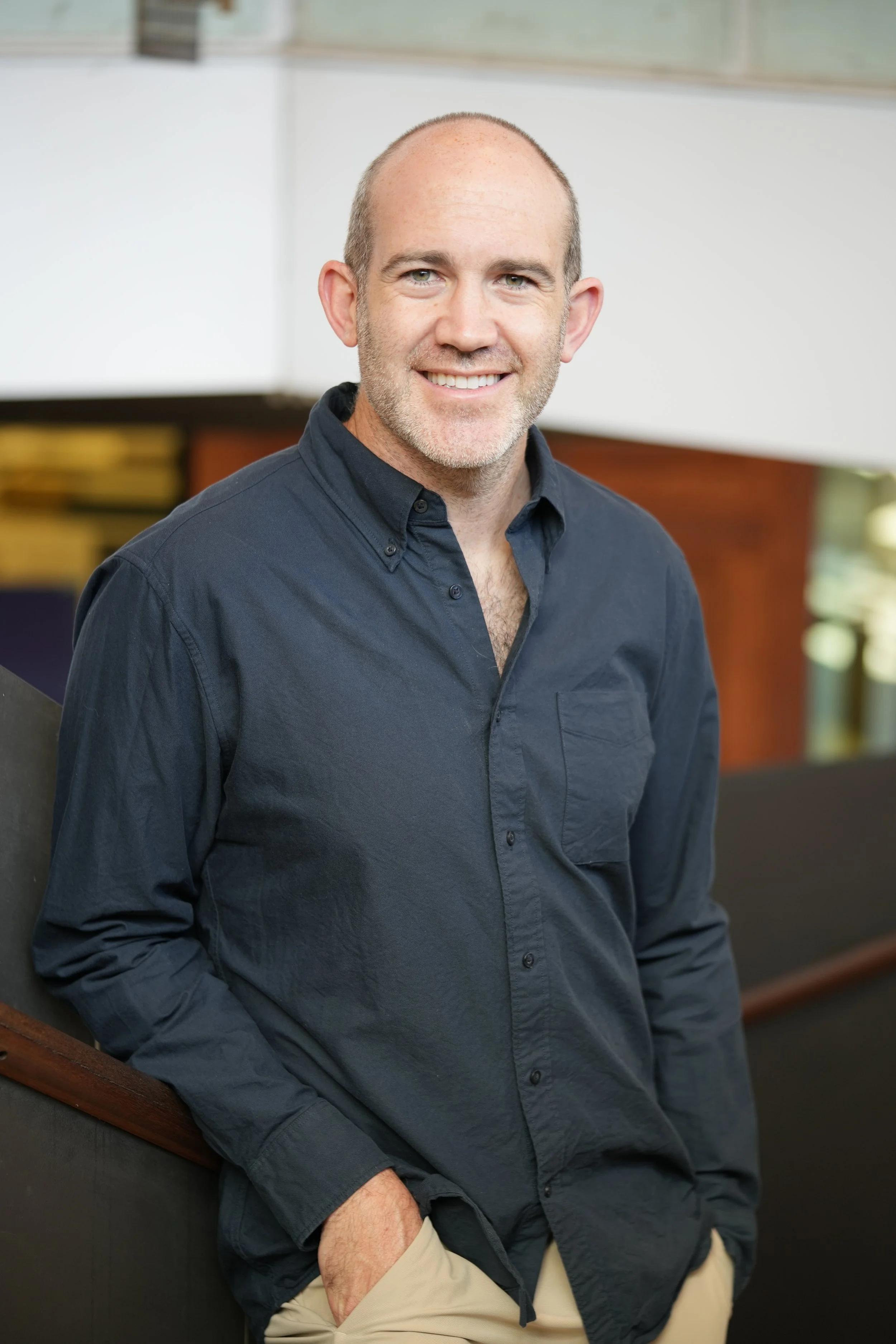 A man with short gray hair and a beard, smiling, wearing a dark blue button-up shirt and beige pants, standing indoors.