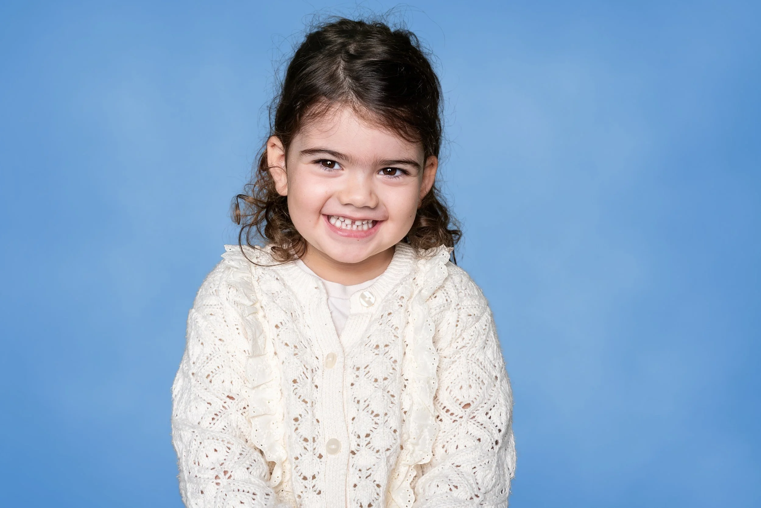 A young girl with dark curly hair, smiling, wearing a cream-colored knit sweater, against a blue background.