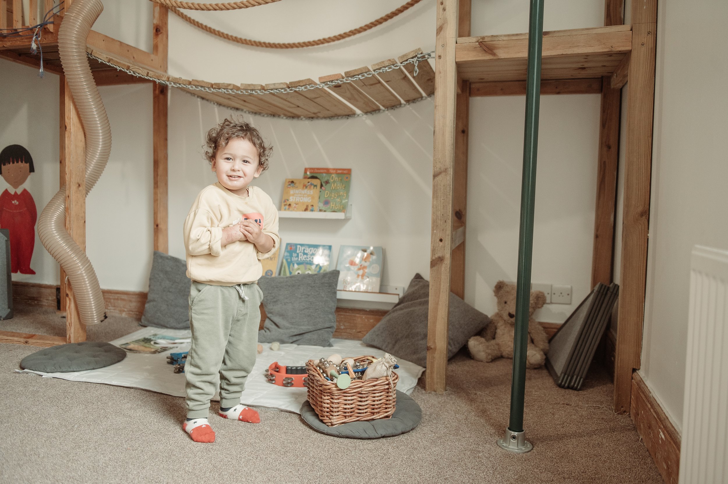 A young boy with curly hair standing in a playroom, smiling, surrounded by cushions, toys, books, and a teddy bear, with a wooden structure and climbing ropes in the background.