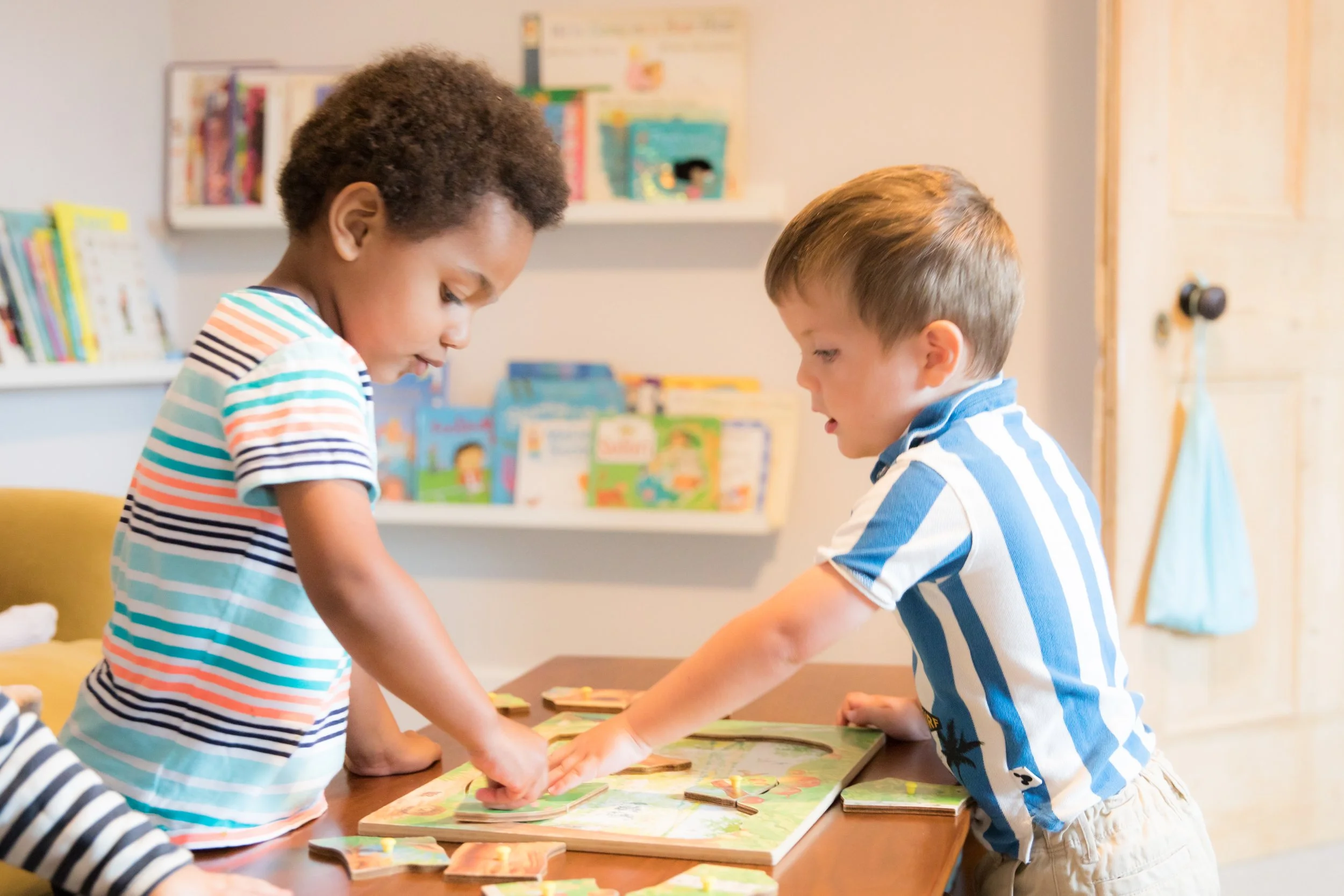 Two young boys are playing with a puzzle on a wooden table in a cozy room. The boy on the left has curly hair and is wearing a striped shirt, while the boy on the right has straight hair and is wearing a blue and white striped shirt. Bookshelves with