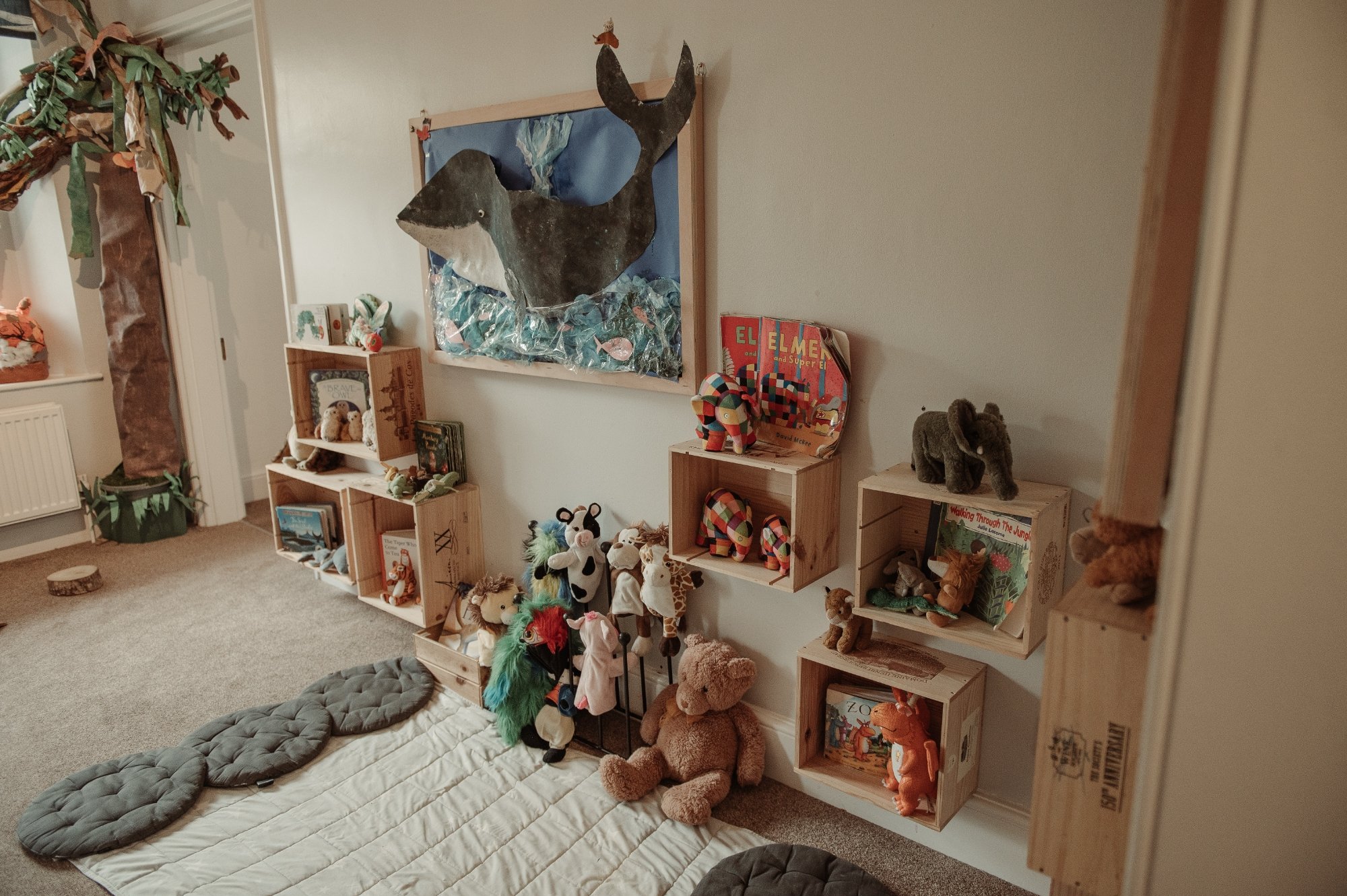 A corner of a nursery with stuffed animals and books on wooden shelves, a large wall art of a whale, and a fabric tree decoration near the window.