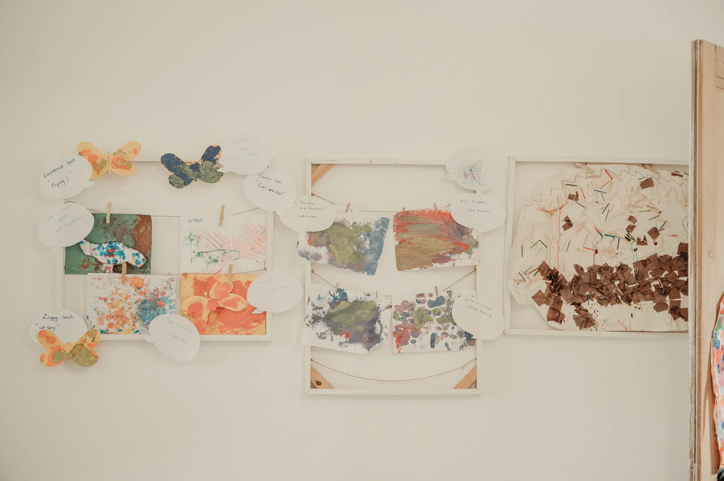Display of children's artwork including paintings, collages, and paper butterflies, with handwritten labels on a white wall.