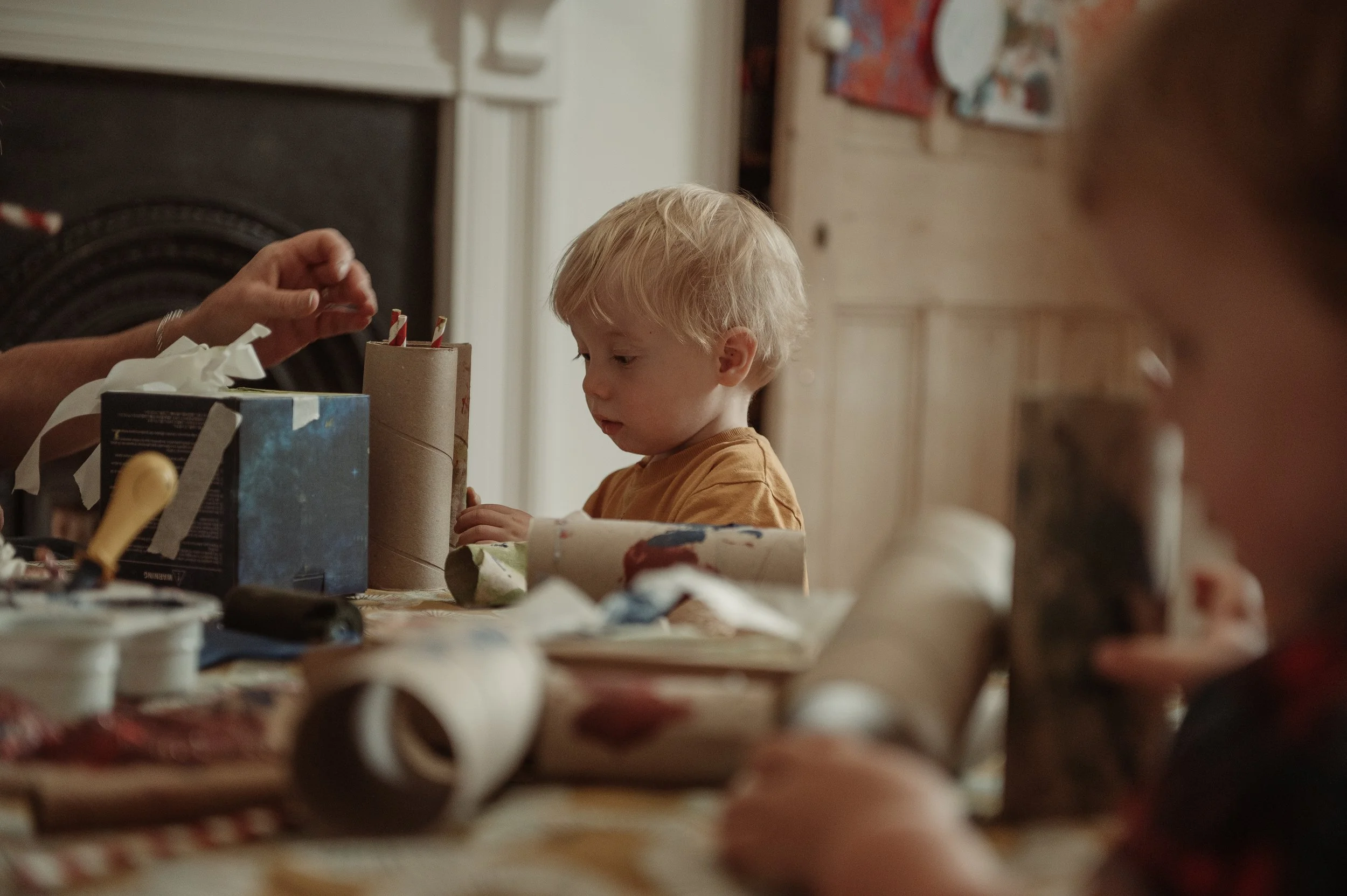 A young blonde boy wearing an orange shirt sitting at a table filled with craft supplies like paper rolls, tape, and a box, concentrating on an activity with an adult's hand assisting him. Another child partially visible on the right side.