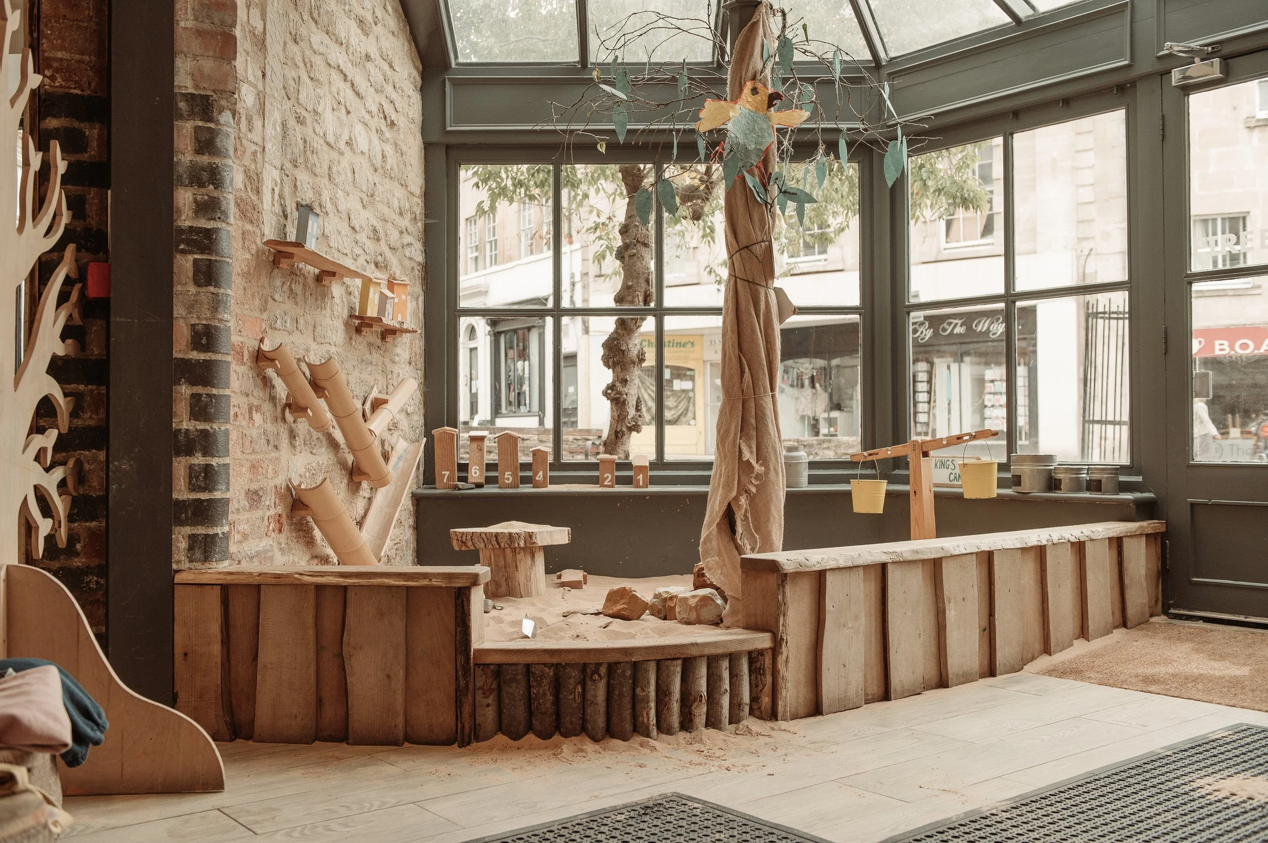 Indoor wooden sandbox play area with natural decor, nearby windows, and outdoor view of shops and street.