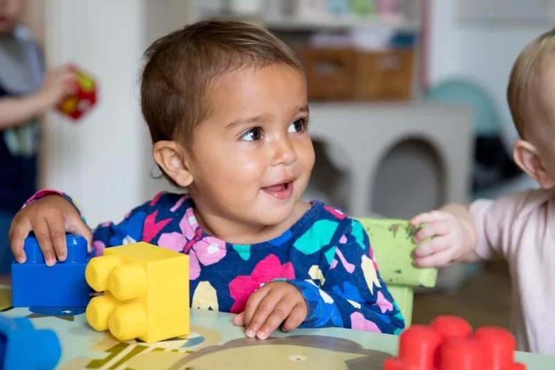 A young girl with short brown hair and a colorful floral shirt sitting at a table with large building blocks, smiling and looking to her right in a brightly lit room.