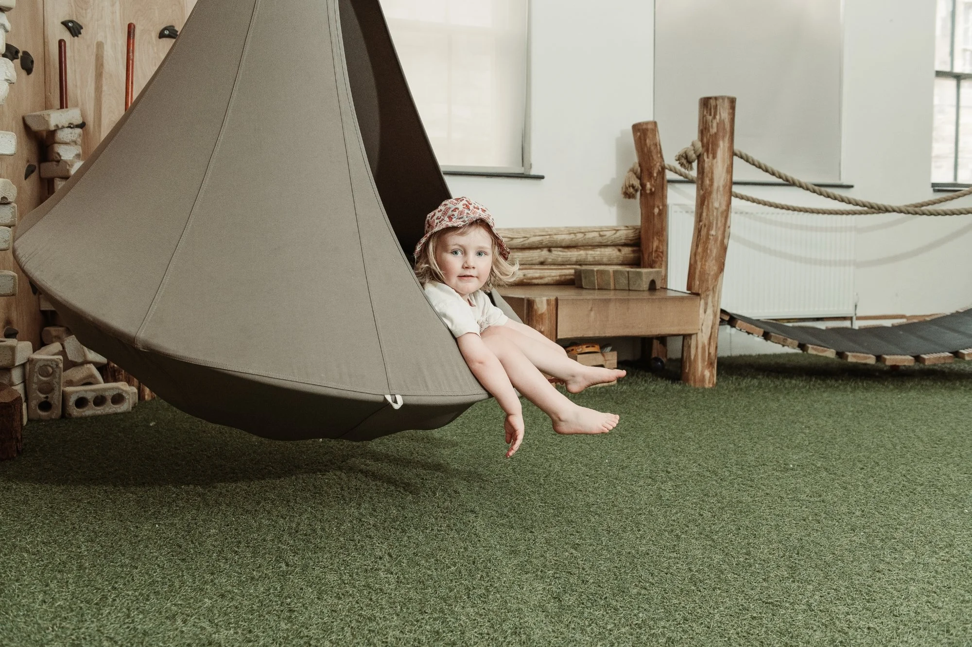 A young girl with blonde hair, wearing a hat and light-colored clothing, sitting inside a large beige fabric hanging chair in an indoor play area. The background features wooden structures, a rope railing, and a small black platform with green grass 