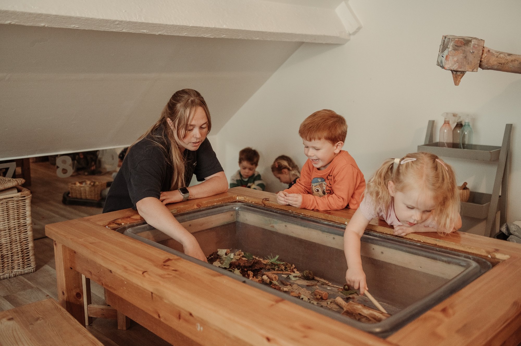 Children and a woman exploring a nature table with logs, leaves, and small natural objects in a cozy indoor setting.
