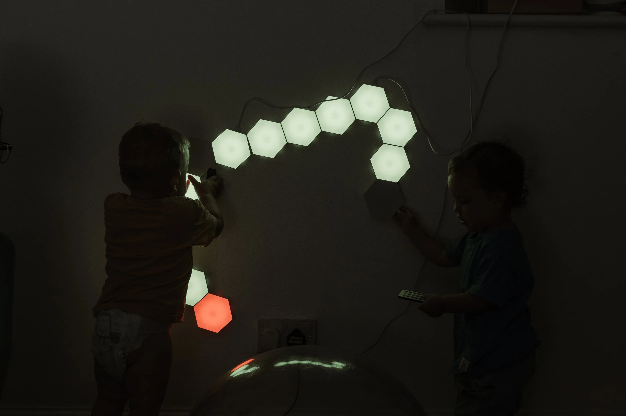 Two children playing with illuminated hexagonal light panels on a wall in a dark room.