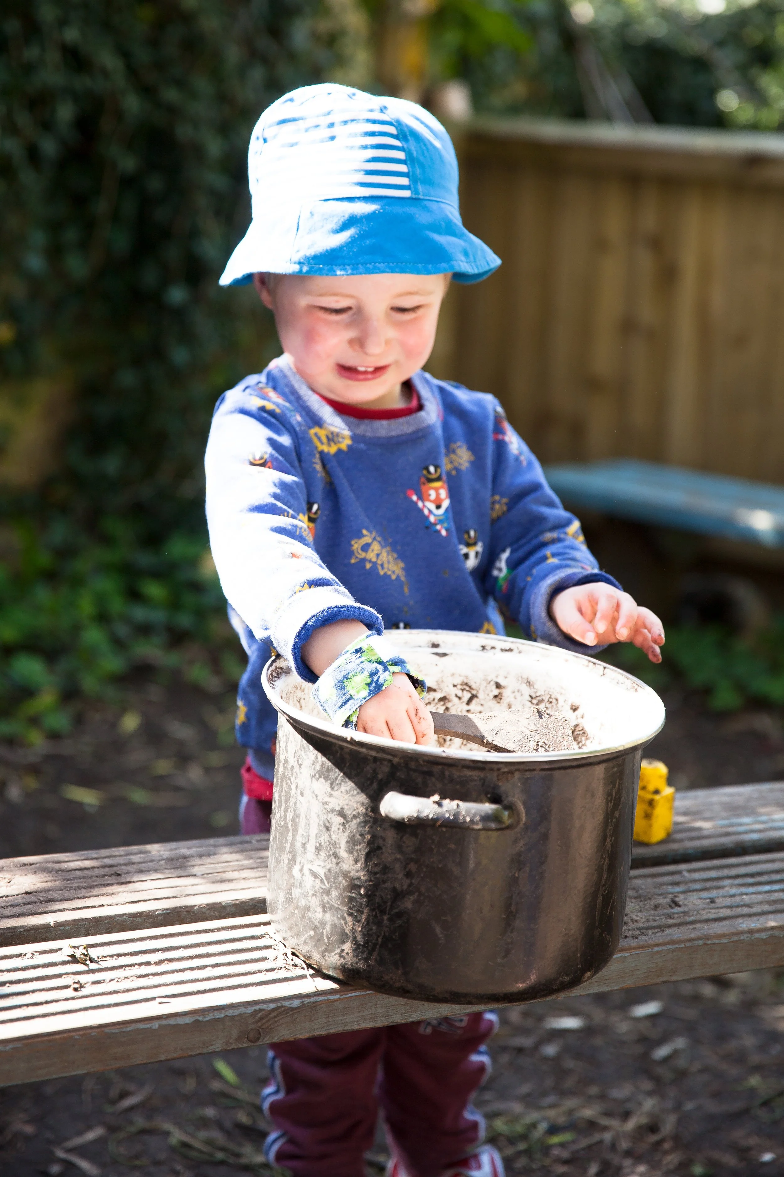 Child wearing a blue bucket hat and a sweater with animal designs, playing with a large pot outdoors on a wooden bench.