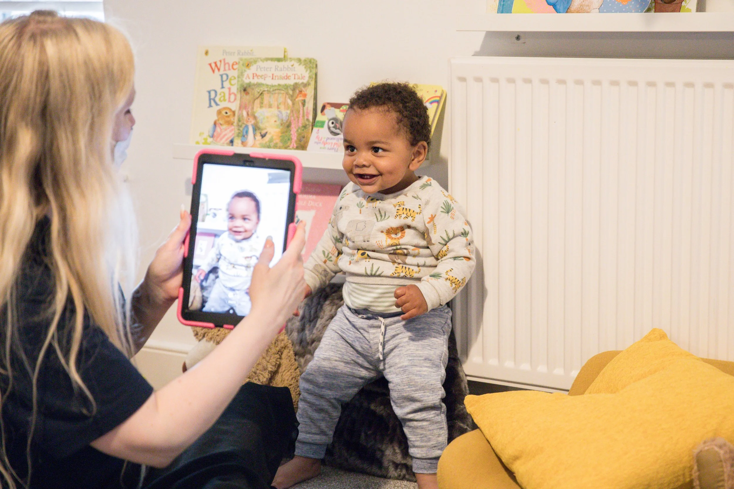 A woman taking a photo of a smiling toddler with a smartphone in a room with books and yellow pillows.