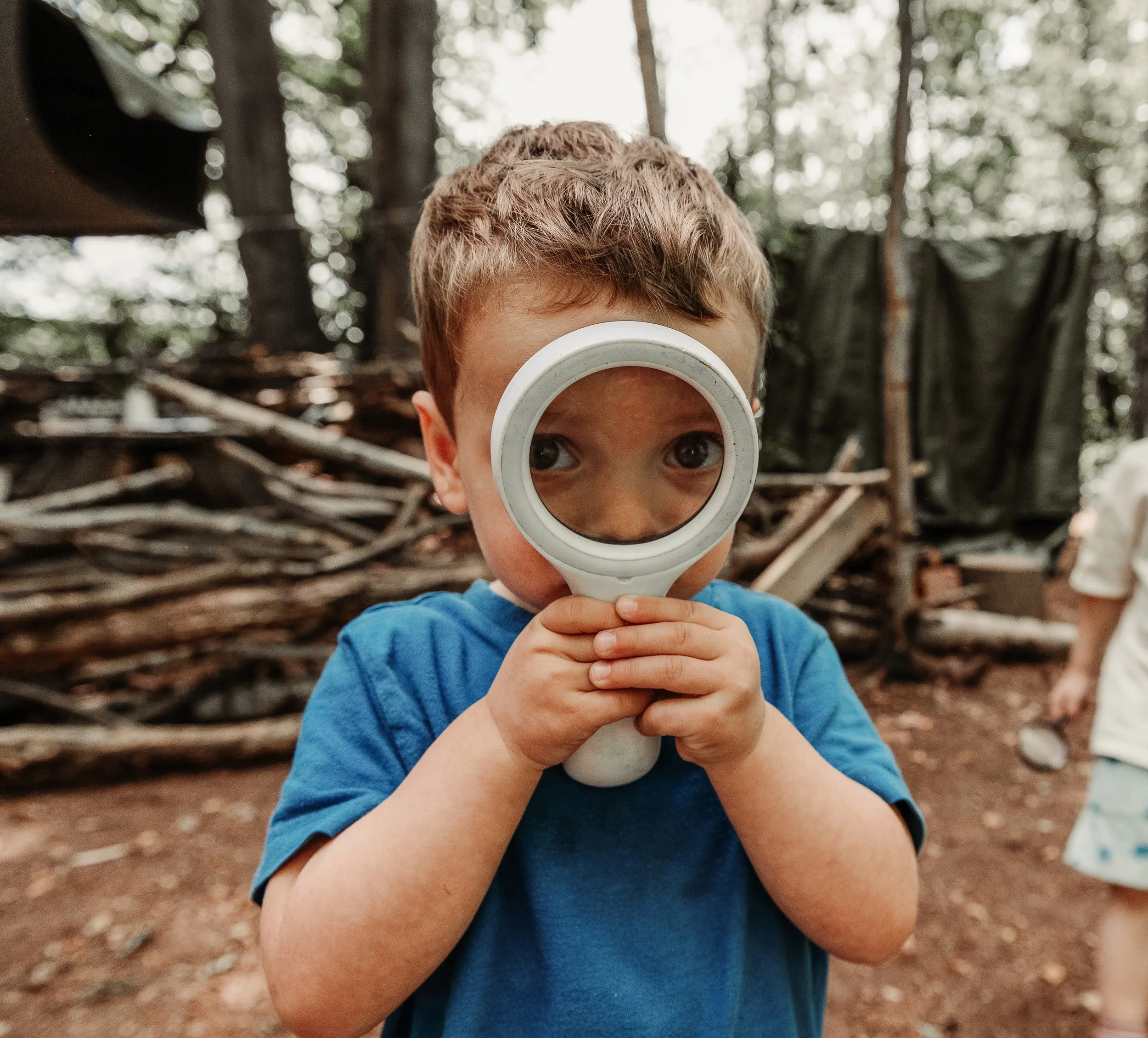 A young boy looking through a magnifying glass in a wooded outdoor setting.