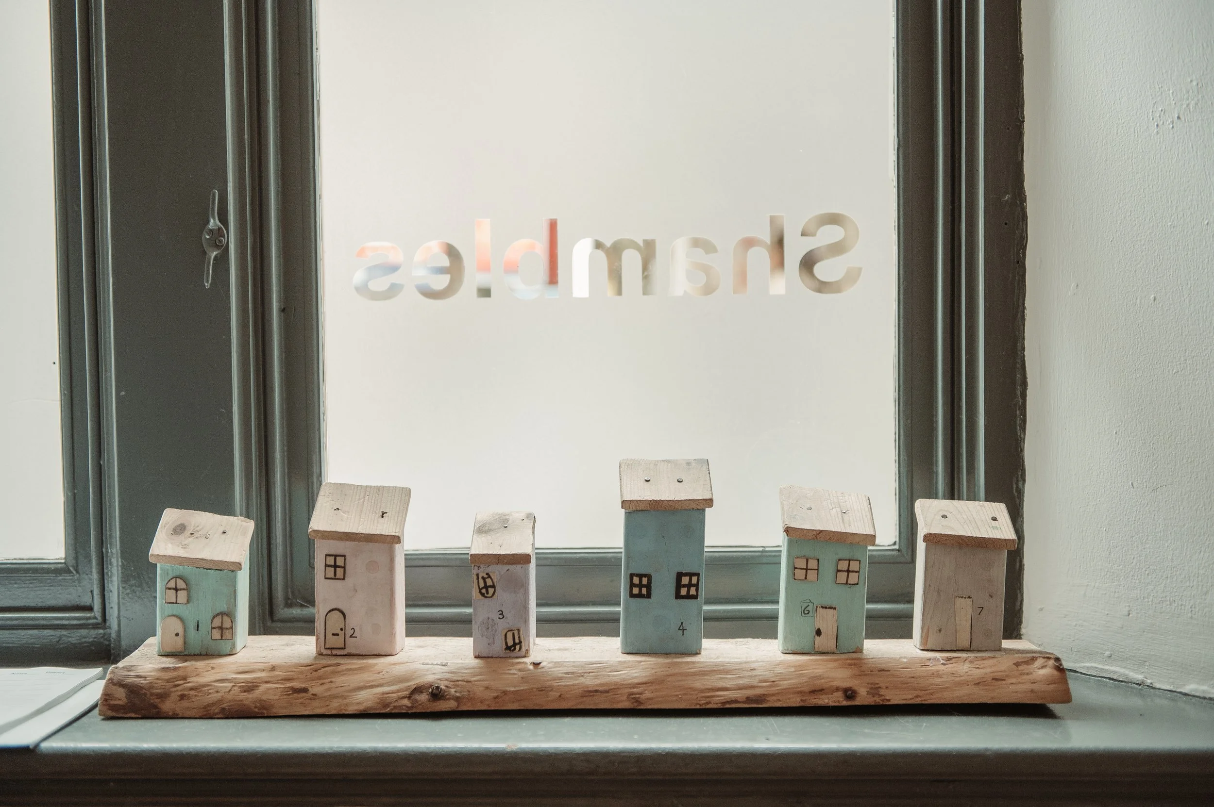 Small decorative houses arranged on a wooden strip on a window sill, with the word 'Shambles' written reversed on the frosted glass window behind them.