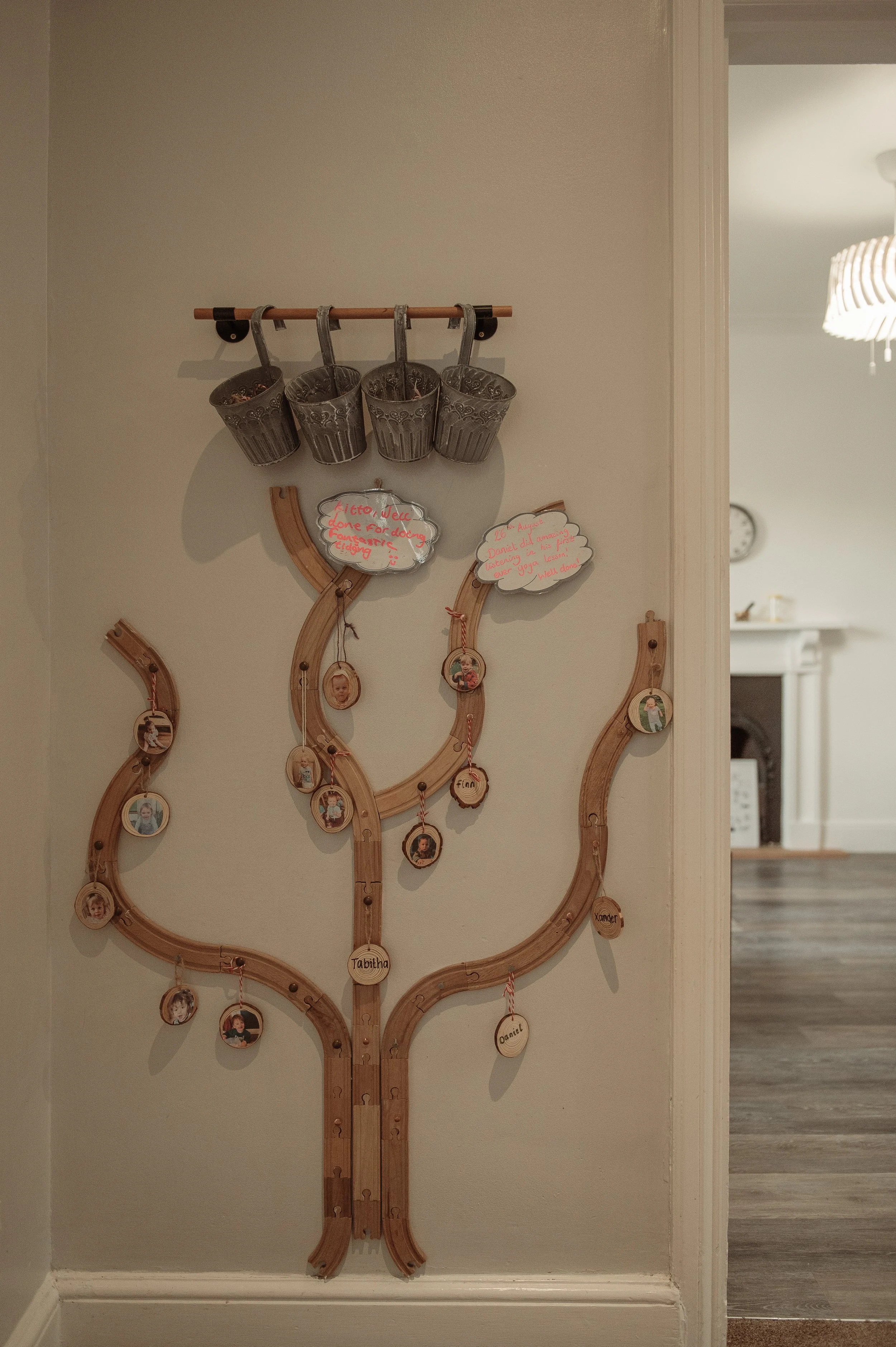 A decorative wooden wall hanging shaped like a tree with small photo frames hanging from its branches, placed on a white wall. Above it, there are four metal cups hanging from a wooden rod.