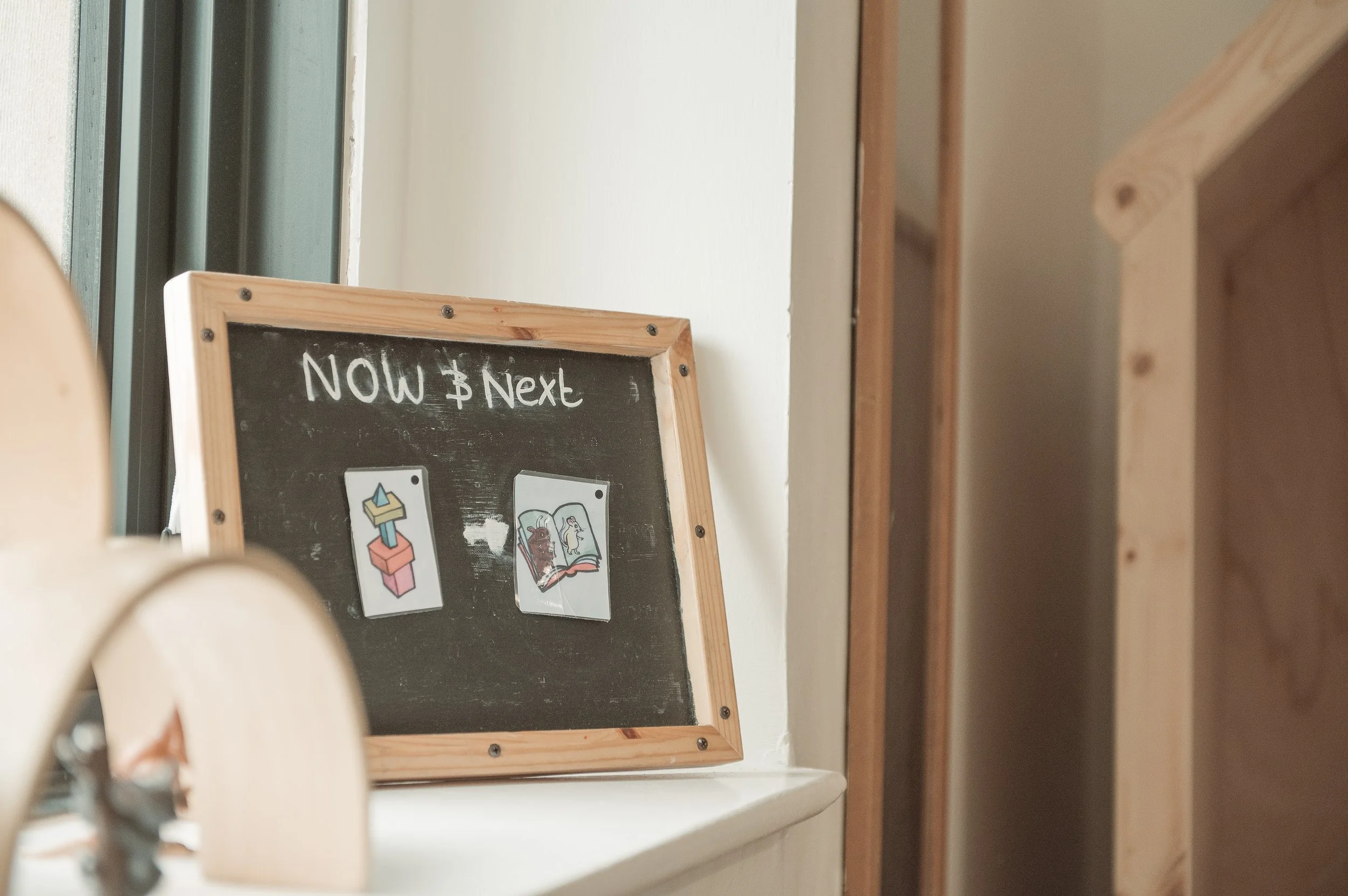 A black chalkboard with a wooden frame on a white windowsill, labeled 'Now & Next', featuring two illustrated cards with images of a toy block and two books.