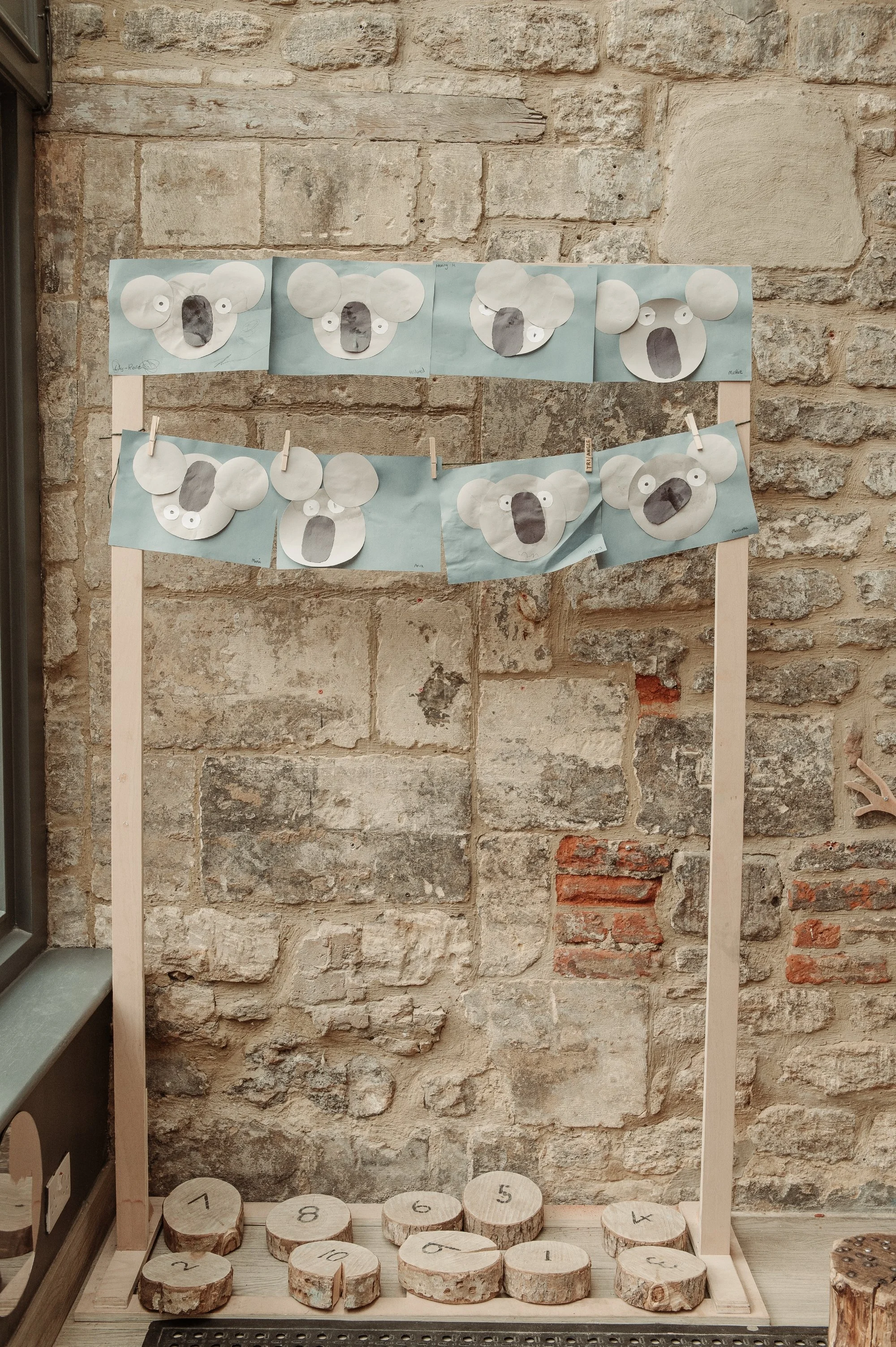 A display of handmade koala paper crafts attached to a string with clothespins, set against a brick wall, with numbered wooden discs at the bottom.