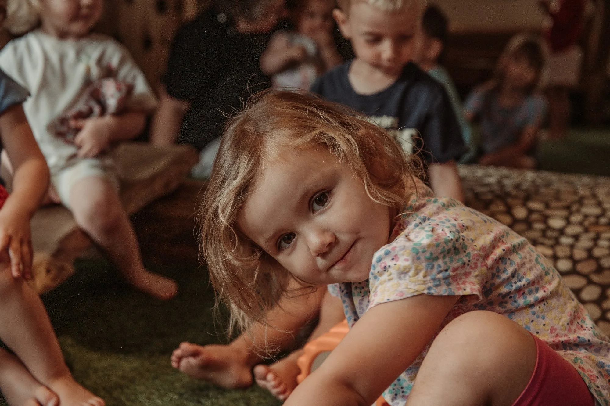 A young girl with curly blond hair leaning forward and smiling at the camera, surrounded by other children sitting on a log and the floor in a cozy, rustic setting.