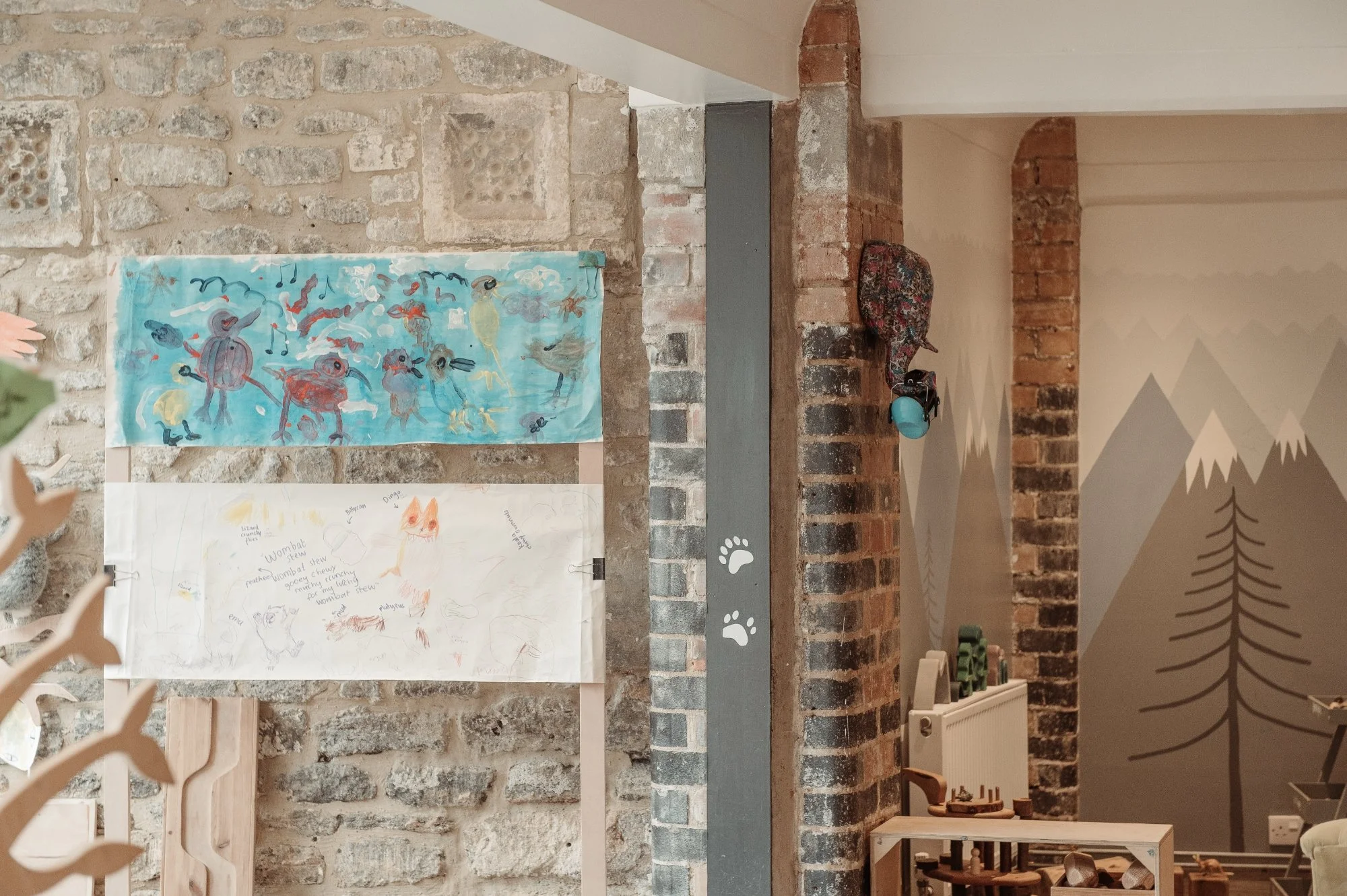 Interior of a cozy room with exposed brick and stone walls, decorated with children's artwork, a wooden shelf, and a mural of mountain peaks.