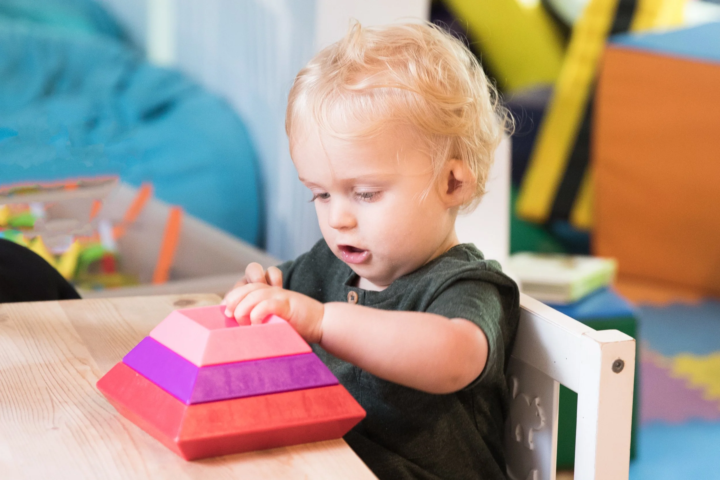 A young child with blonde curly hair playing with stacking toys at a wooden table.