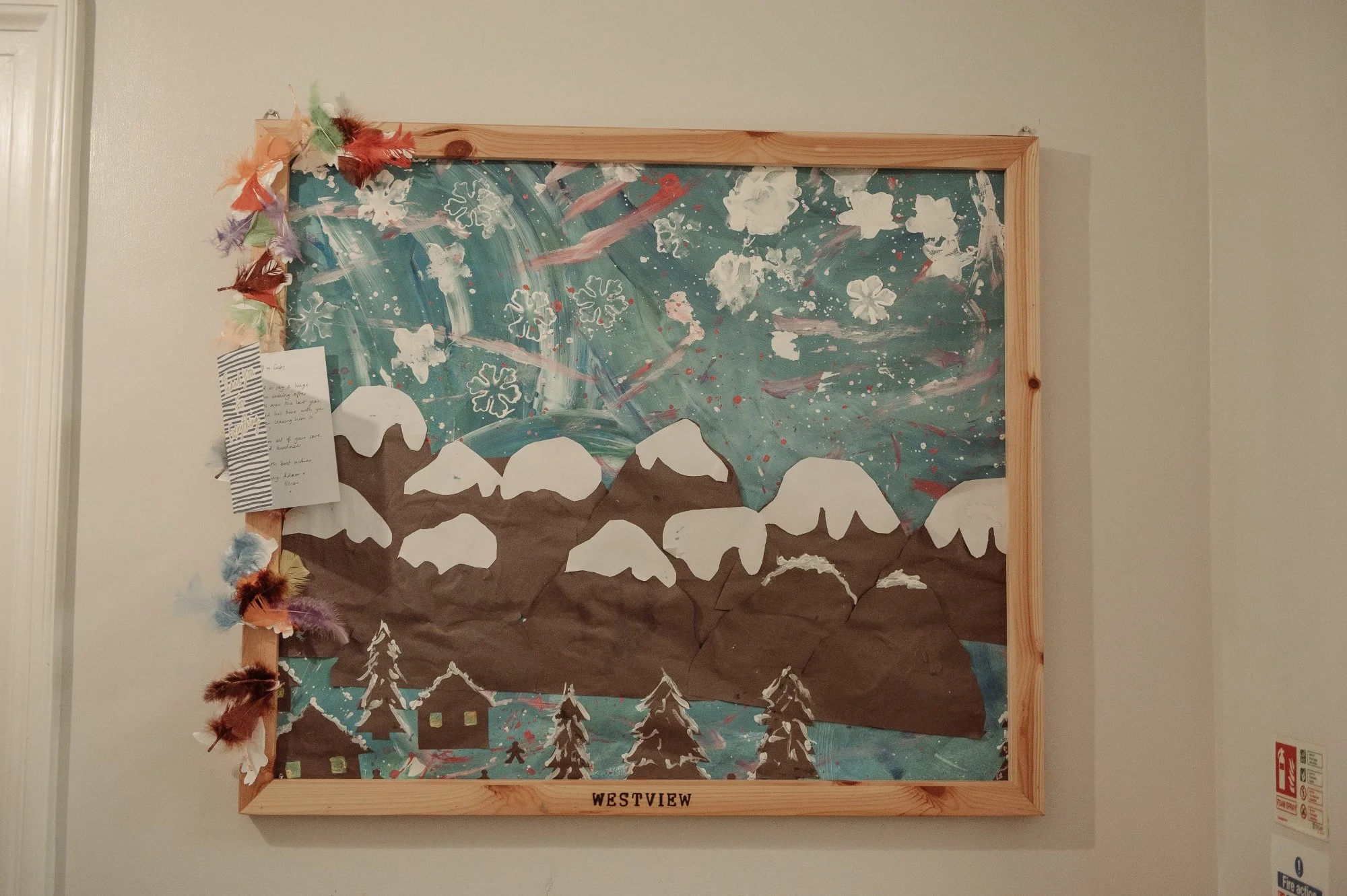 Child's winter mountain scene craft on a wooden frame wall display, featuring mountains with snow, pine trees, and a painted sky with snowflakes, decorated with multicolored feathers and a handwritten note.