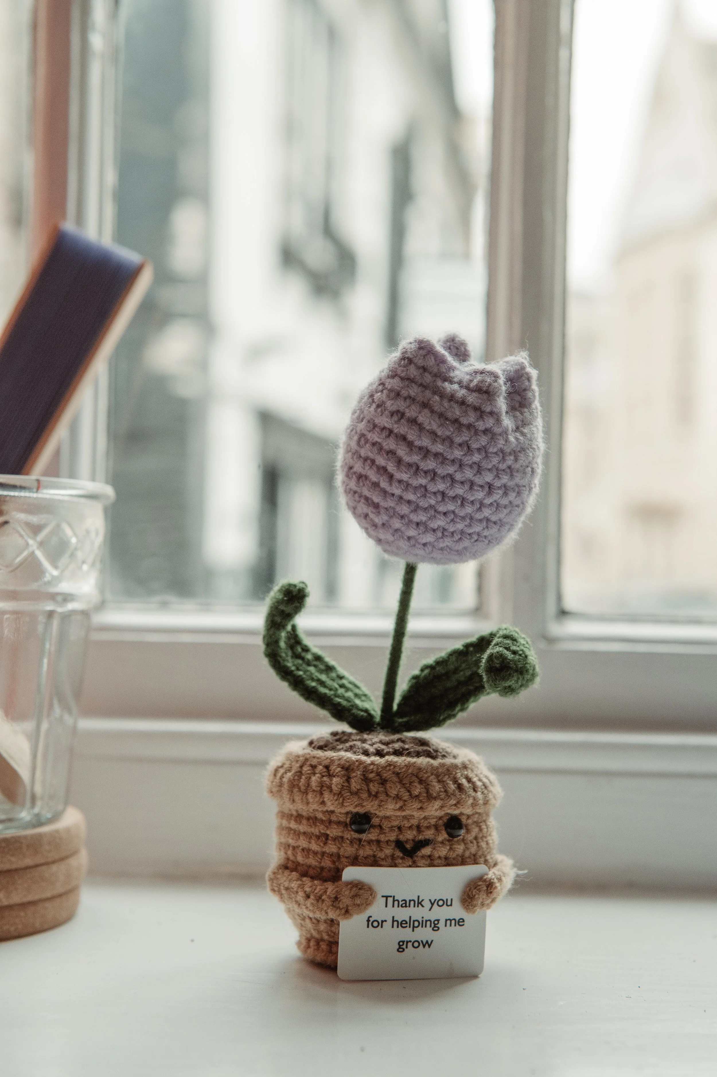 A knitted plush flower with a purple blossom and green leaves in a beige pot with a smiling face, holding a small white sign that says, 'Thank you for helping me grow,' sitting near a window.