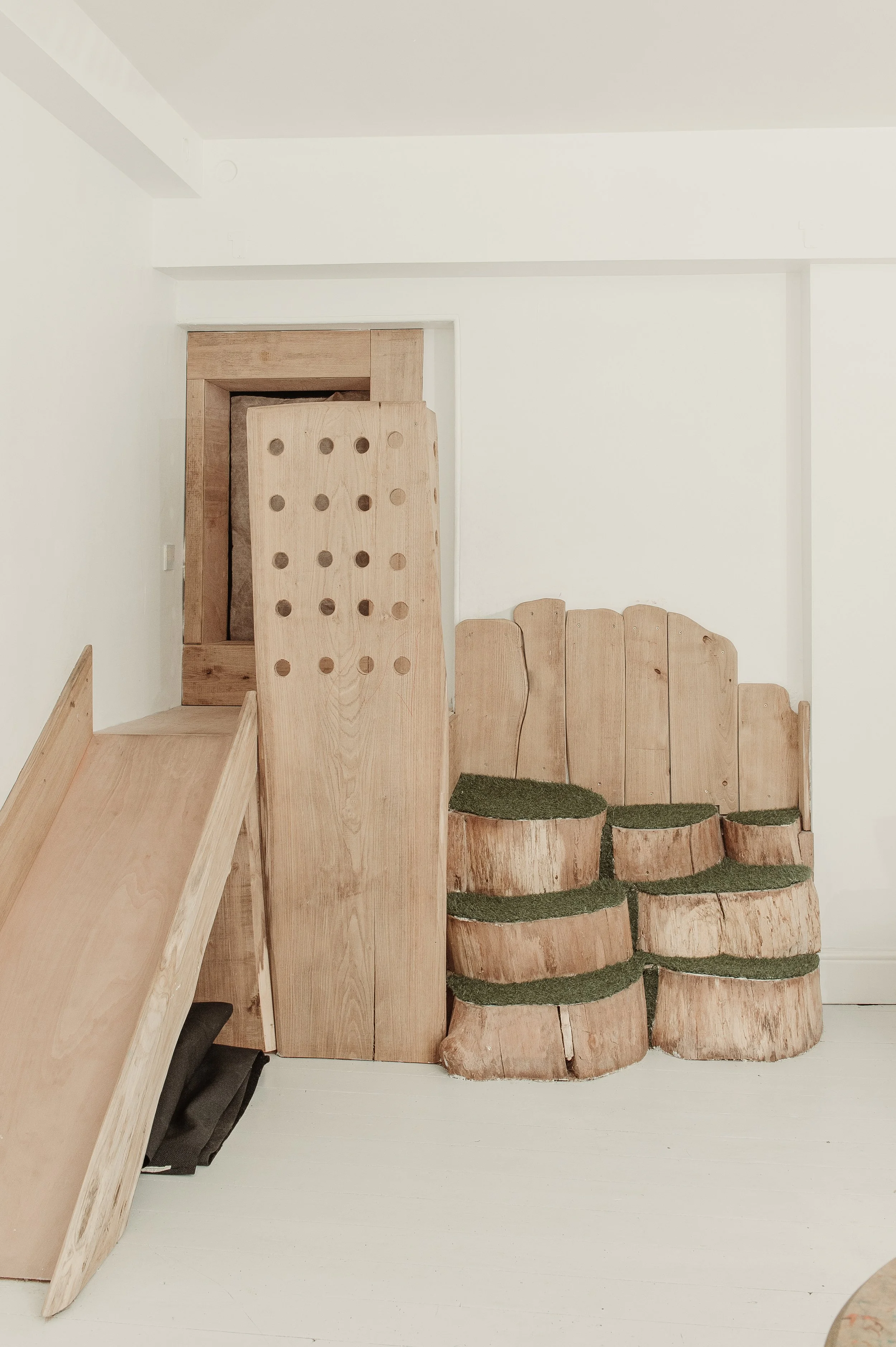 Stacked wooden furniture and decorations, including tree stump seats with green cushioning, against a plain white wall.