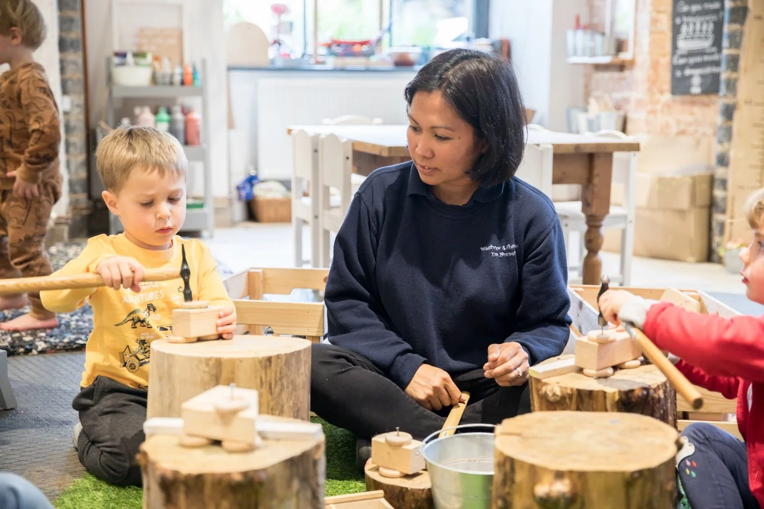 A woman and two young children engaging in a woodworking activity in a well-lit indoor space. The children are using small hammers on wood blocks, supervised by the woman.