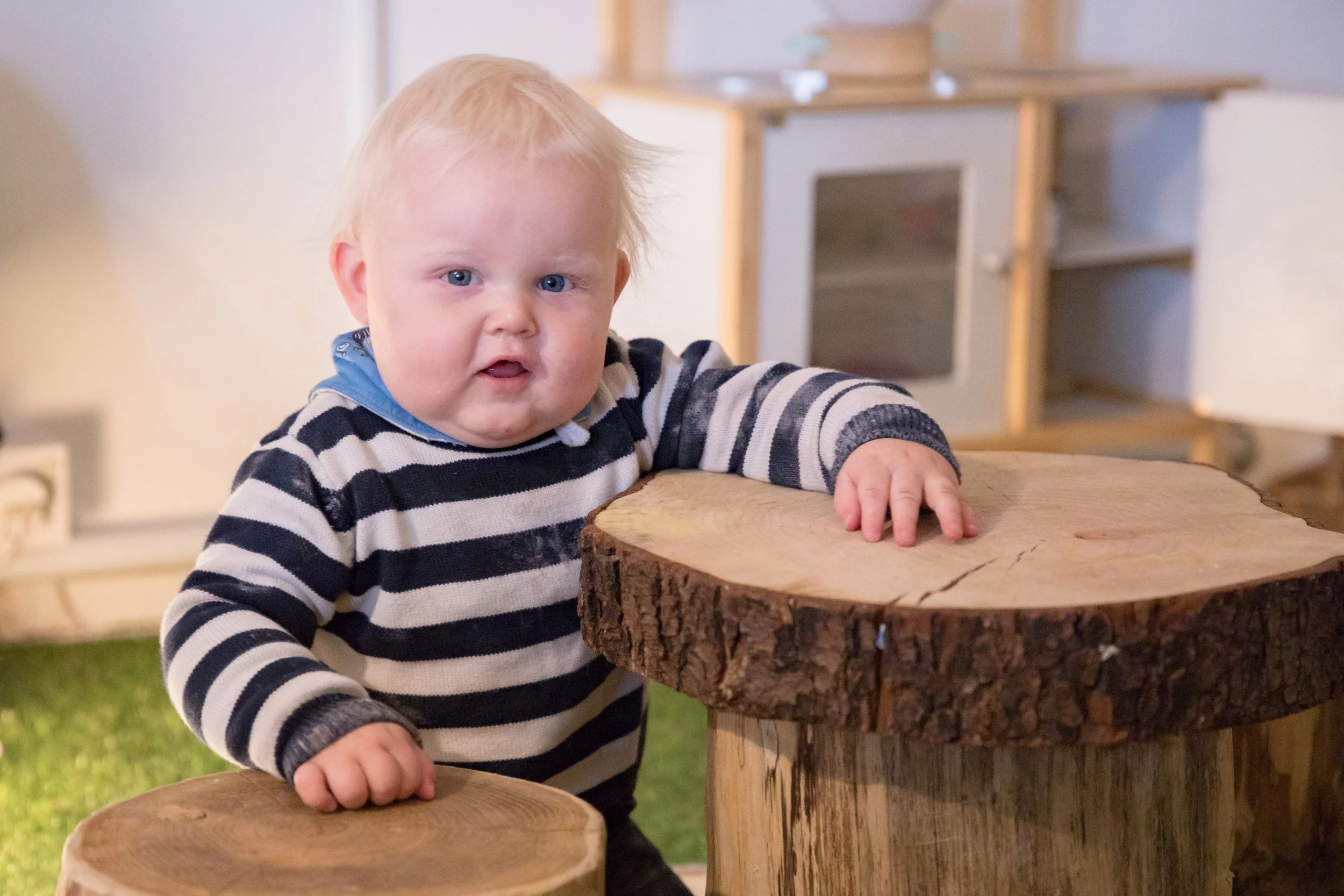 A young child with blonde hair and blue eyes, wearing a black and white striped sweater, pressing one hand on a large wooden log used as a table, in an indoor setting.