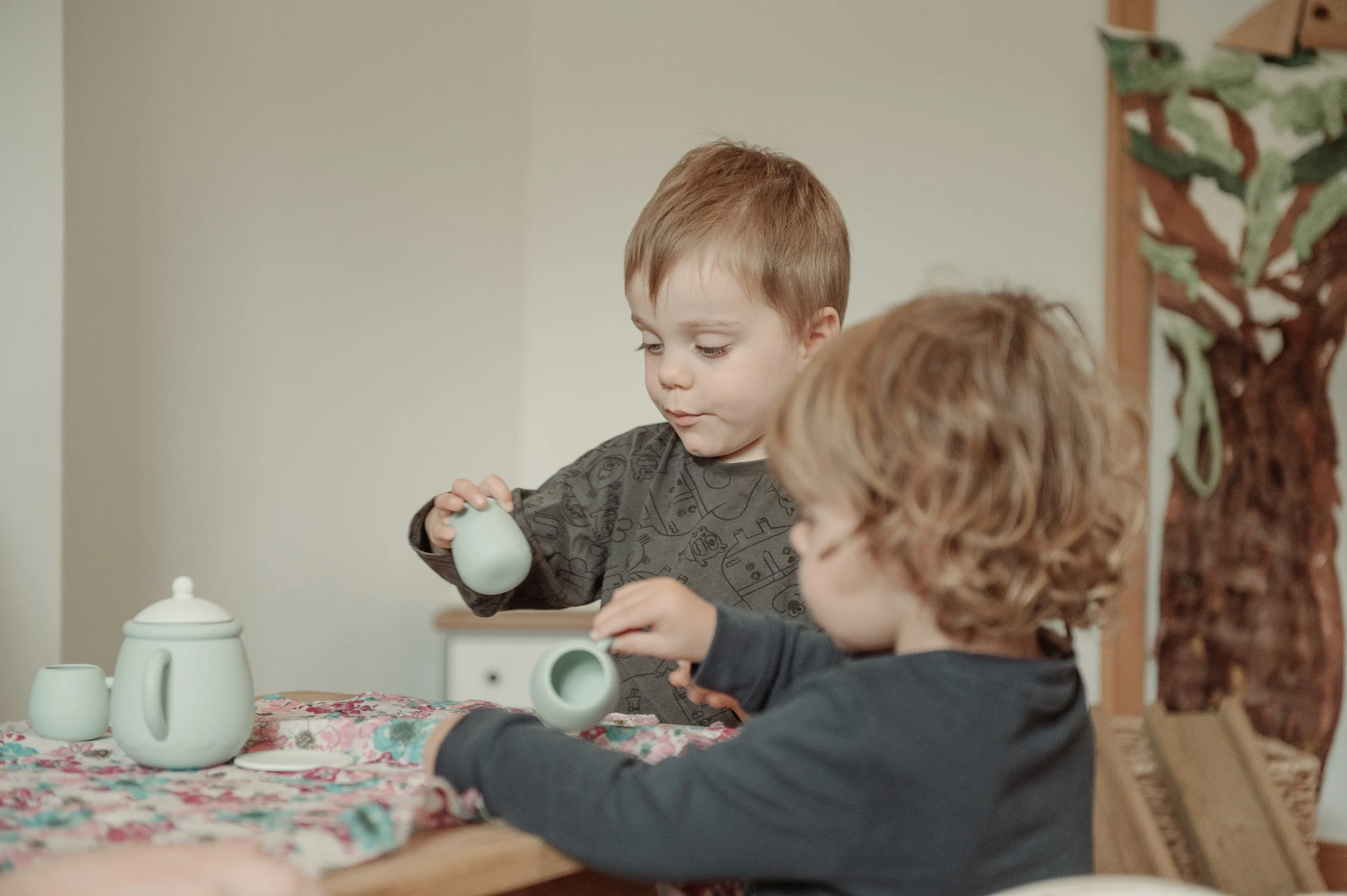 Two young children with light brown hair playing with a teal ceramic tea set on a floral tablecloth at a wooden table.