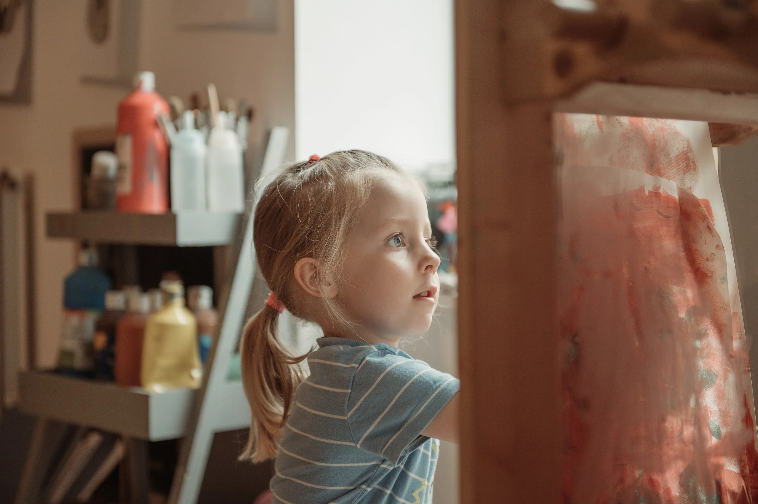 A young girl with blonde hair in pigtails, wearing a striped shirt, is looking at something off-camera in an art studio or classroom. There are art supplies, including paint bottles, on shelves in the background.