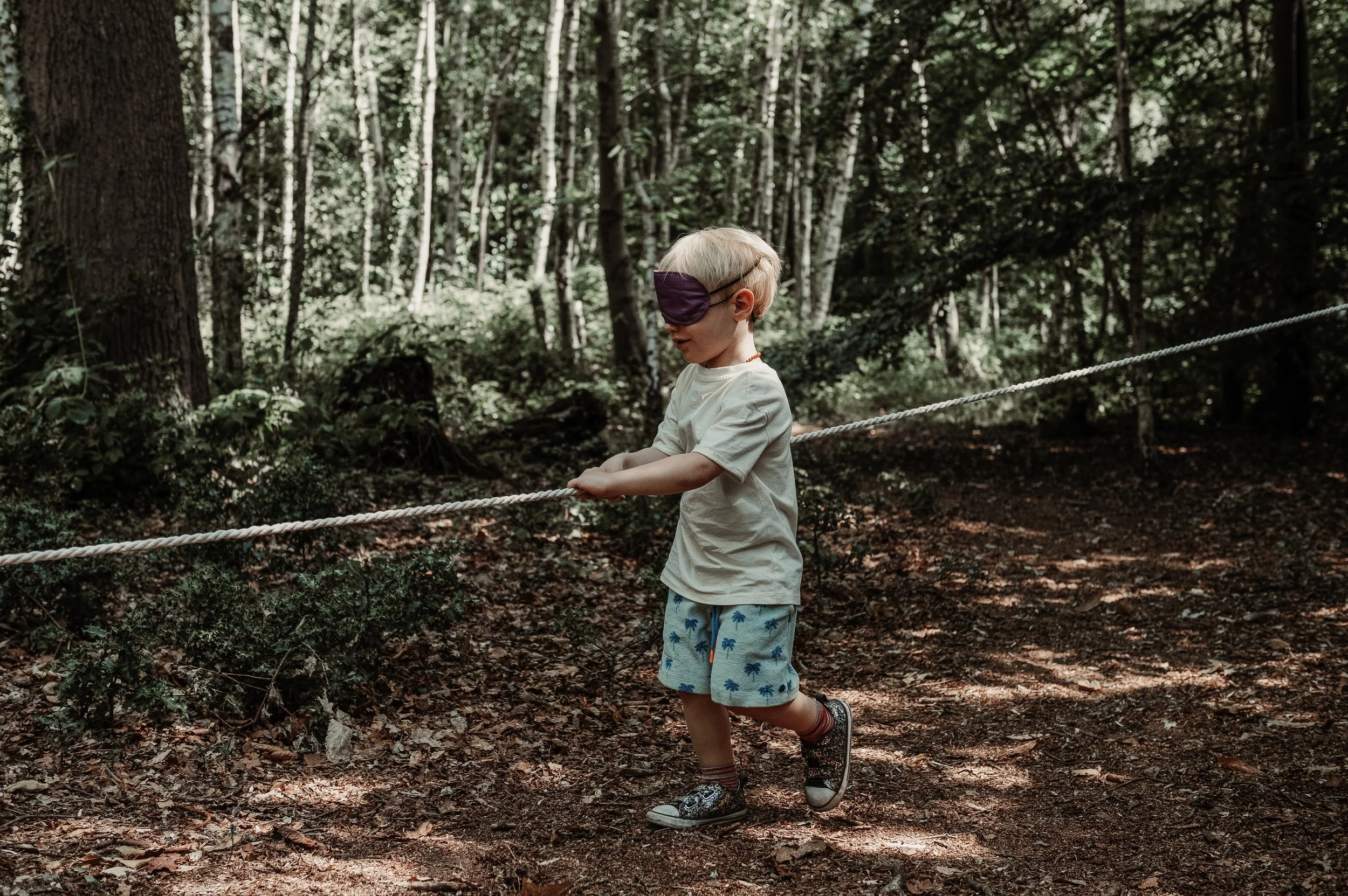 A young boy with blonde hair, wearing a blindfold, runnable shorts with palm tree prints, a beige t-shirt, and sneakers, holding a rope in a forest setting.