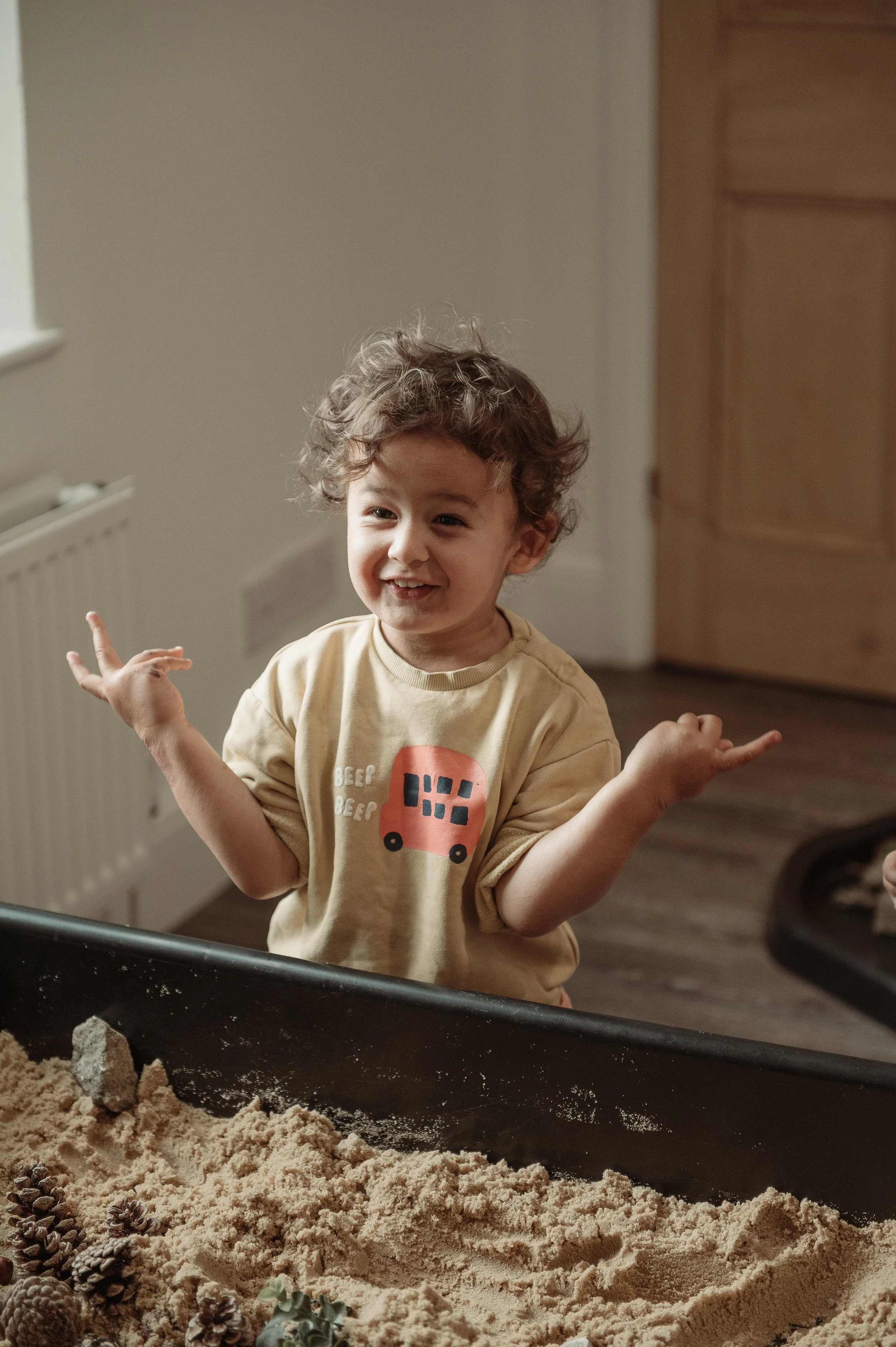 A young boy with curly hair smiling and gesturing with his hands near a sandbox filled with sand, small rocks, and pinecones inside a room.