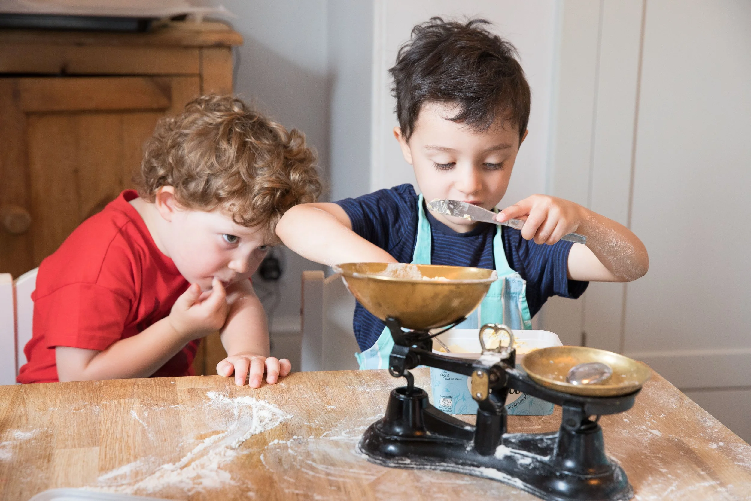 Two young boys baking in a kitchen, one measuring ingredients with a spoon and the other watching closely, with flour scattered on the wooden table.