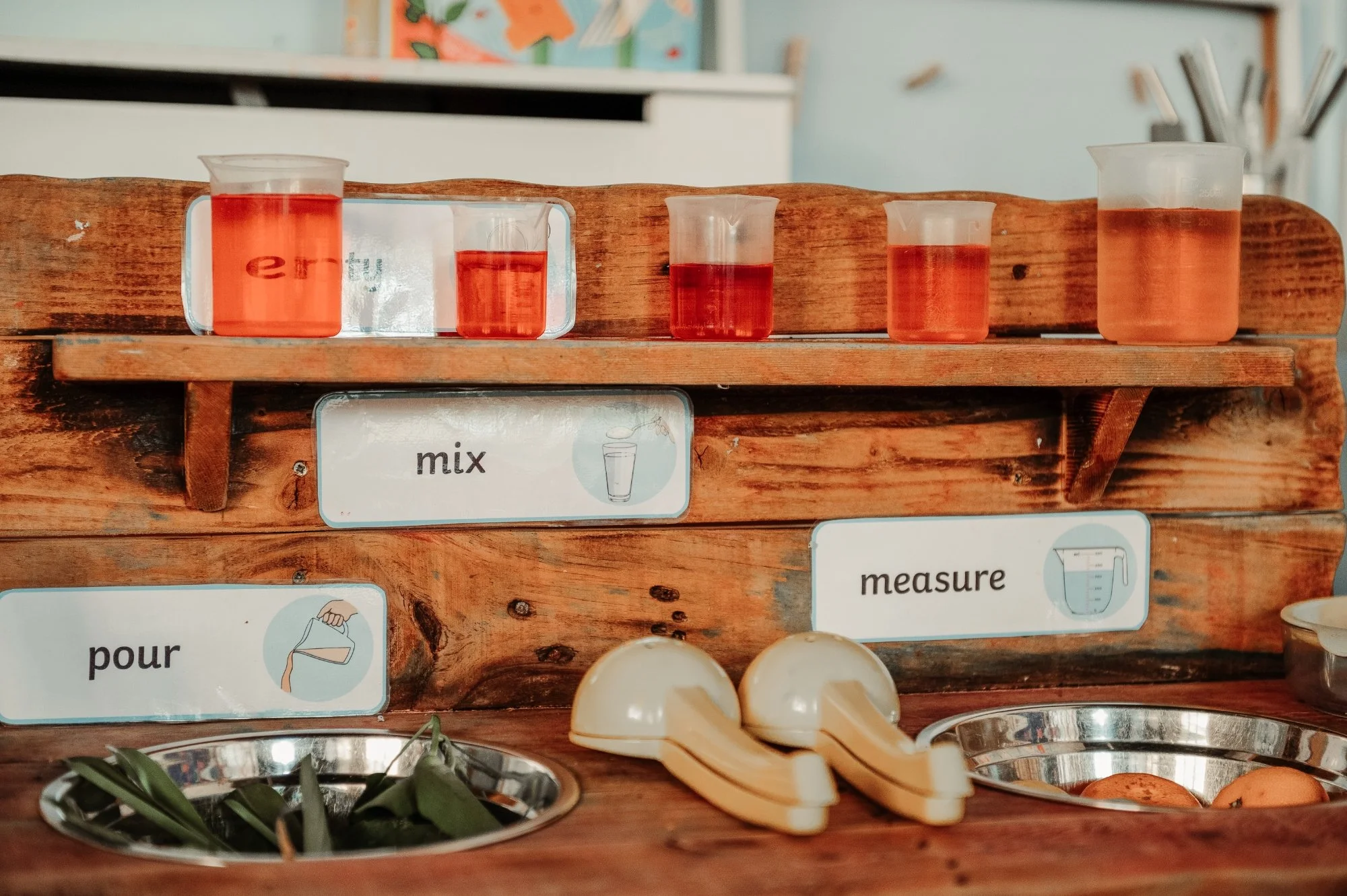 A wooden kitchen counter with four small plastic containers filled with a pink liquid, labeled with adding directions such as 'pour,' 'mix,' and 'measure.' There are two small yellow hand mixers, two silver bowls with green herbs, and a bowl with egg