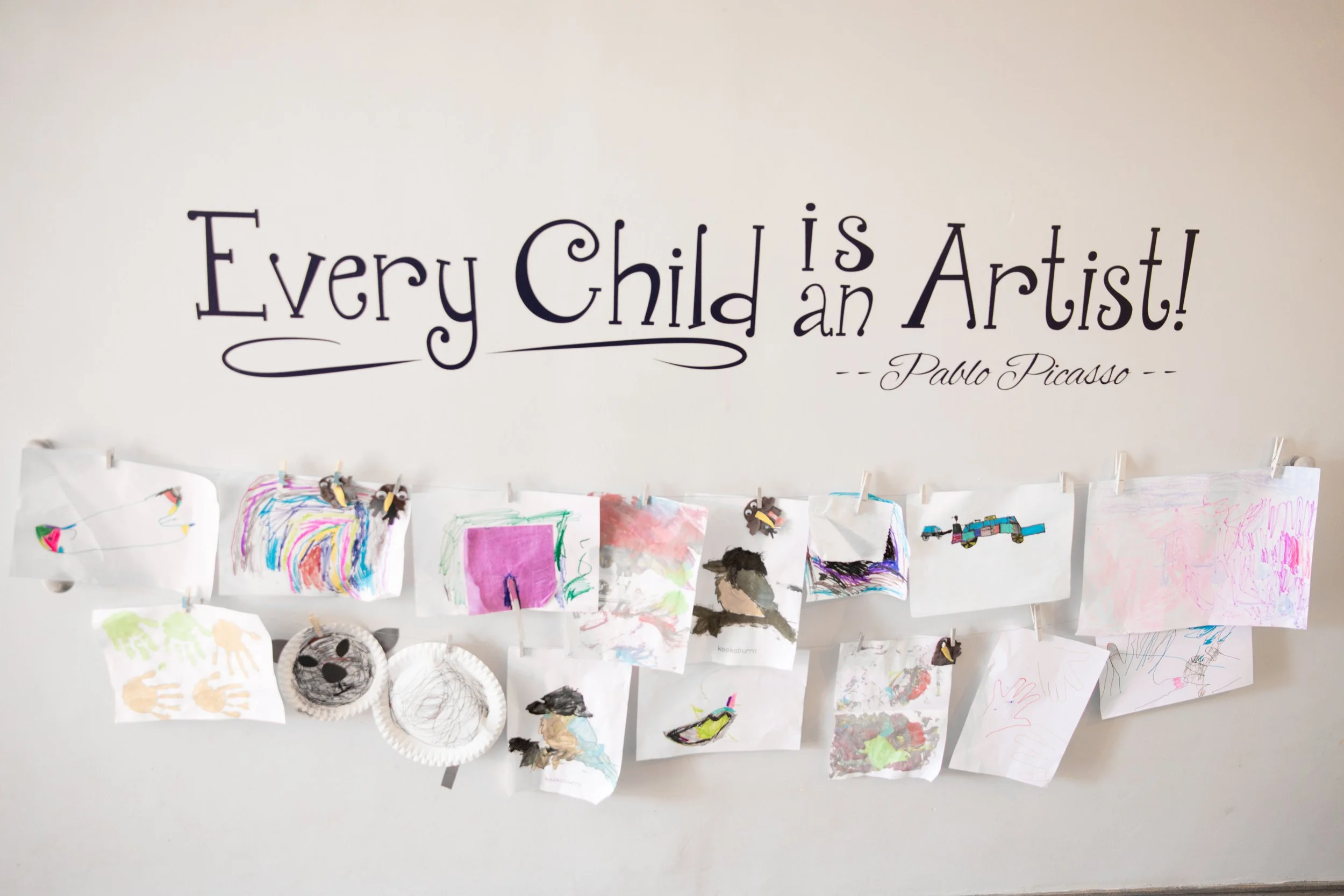 Children's colorful drawings and crafts hanging on a wire wall display with a quote on the wall that reads, 'Every Child is an Artist!' attributed to Pablo Picasso.