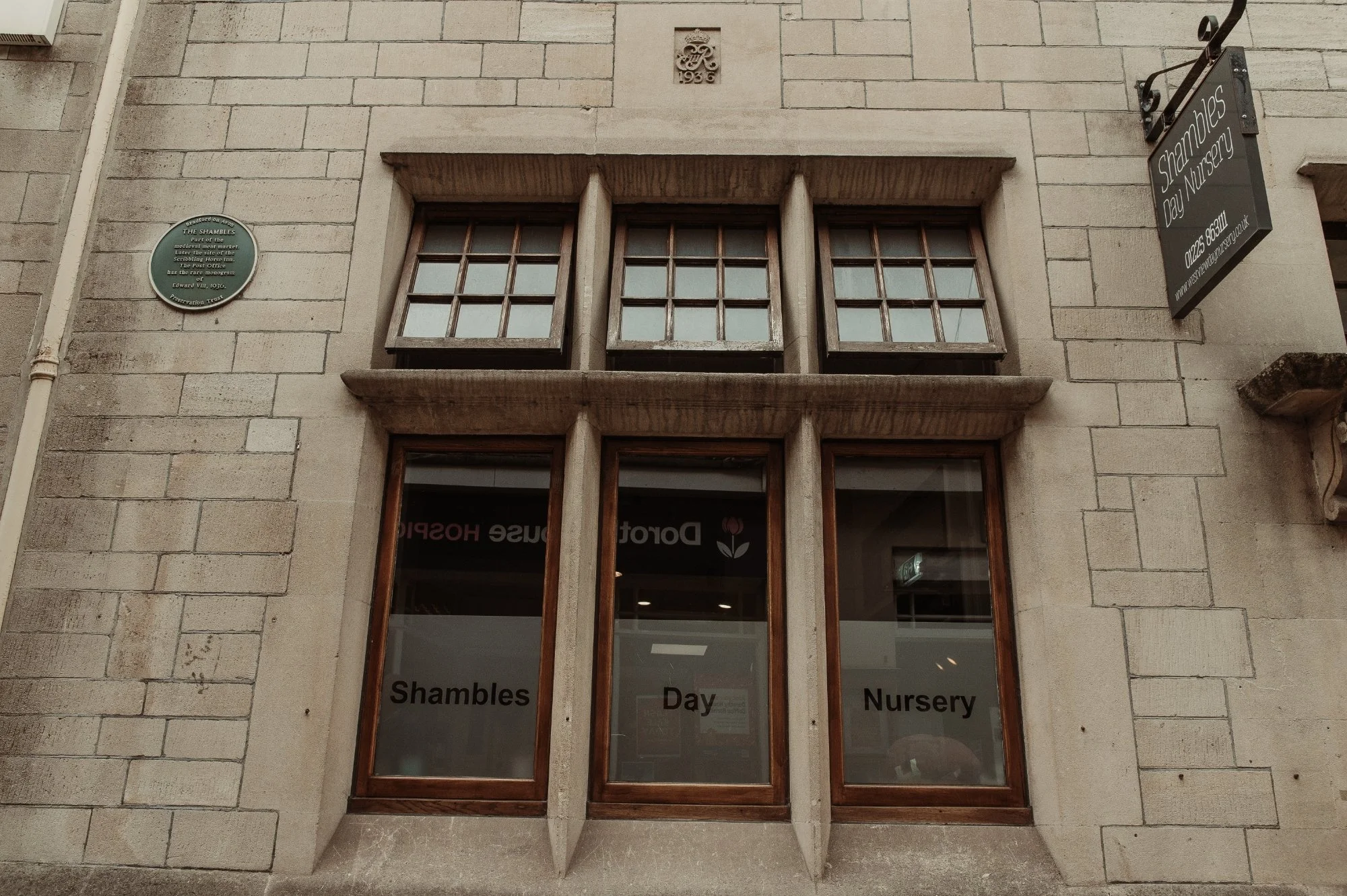 The exterior of a building on The Shambles in Bradford on Avon with a sign that reads 'Shambles Day Nursery' on the right side and windows with the same name on the glass. The building has a stone facade with a plaque and a crest above the windows.